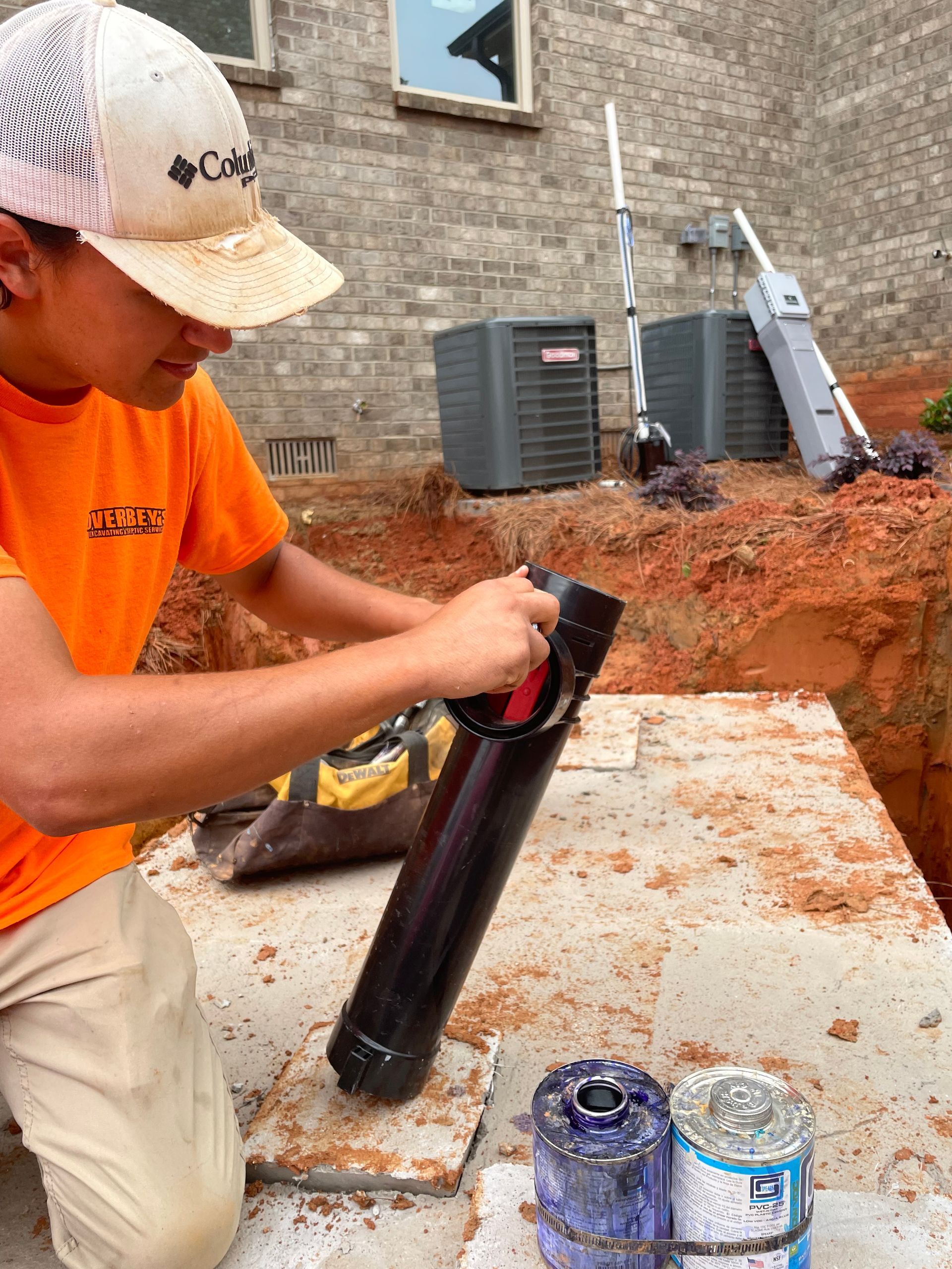 Man in orange shirt cutting black pipe in a construction site, with tools and air conditioning units in the background.