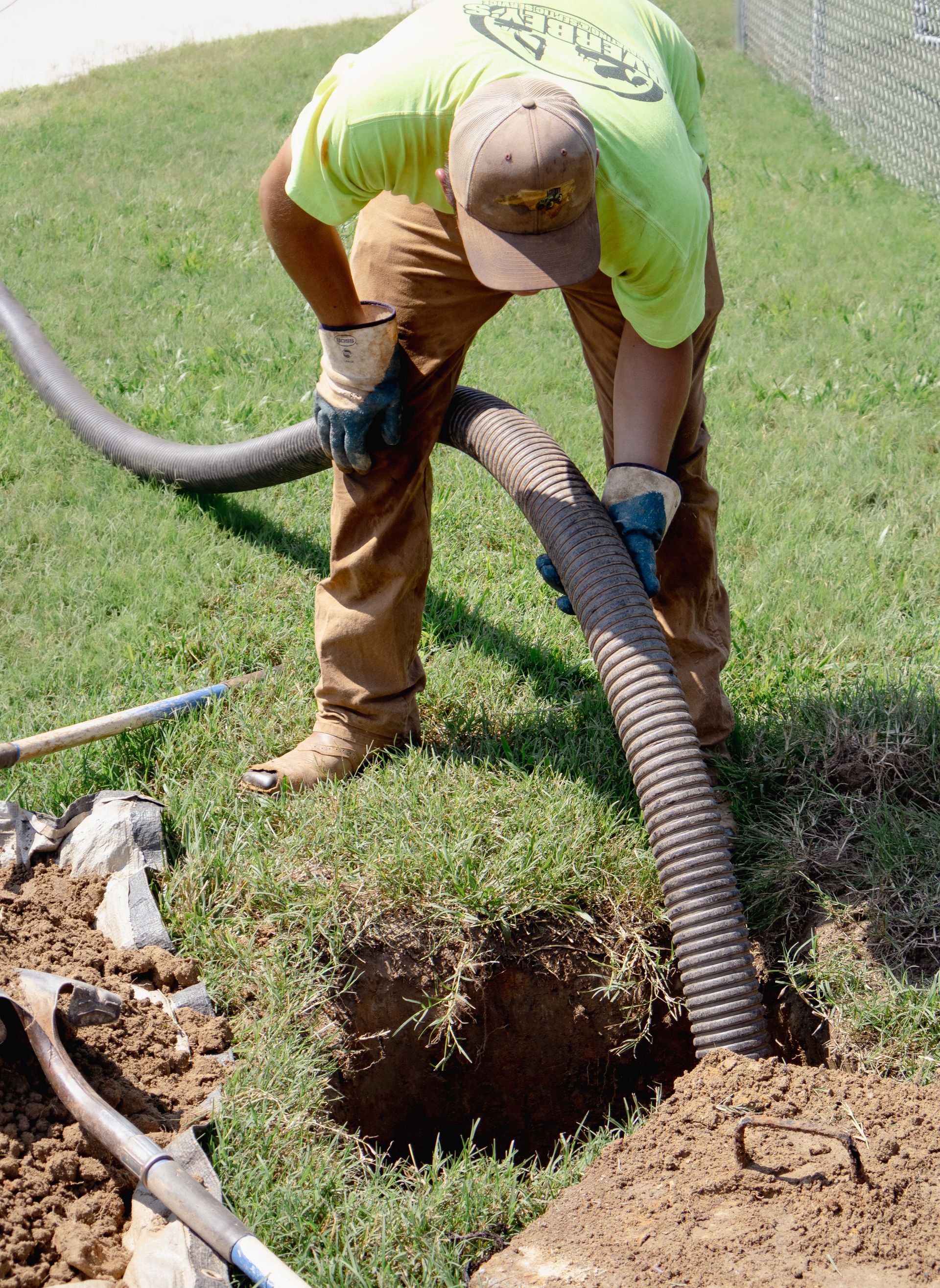 Man in safety gear working in a hole, holding a large hose. Brown dirt and green grass surround him.