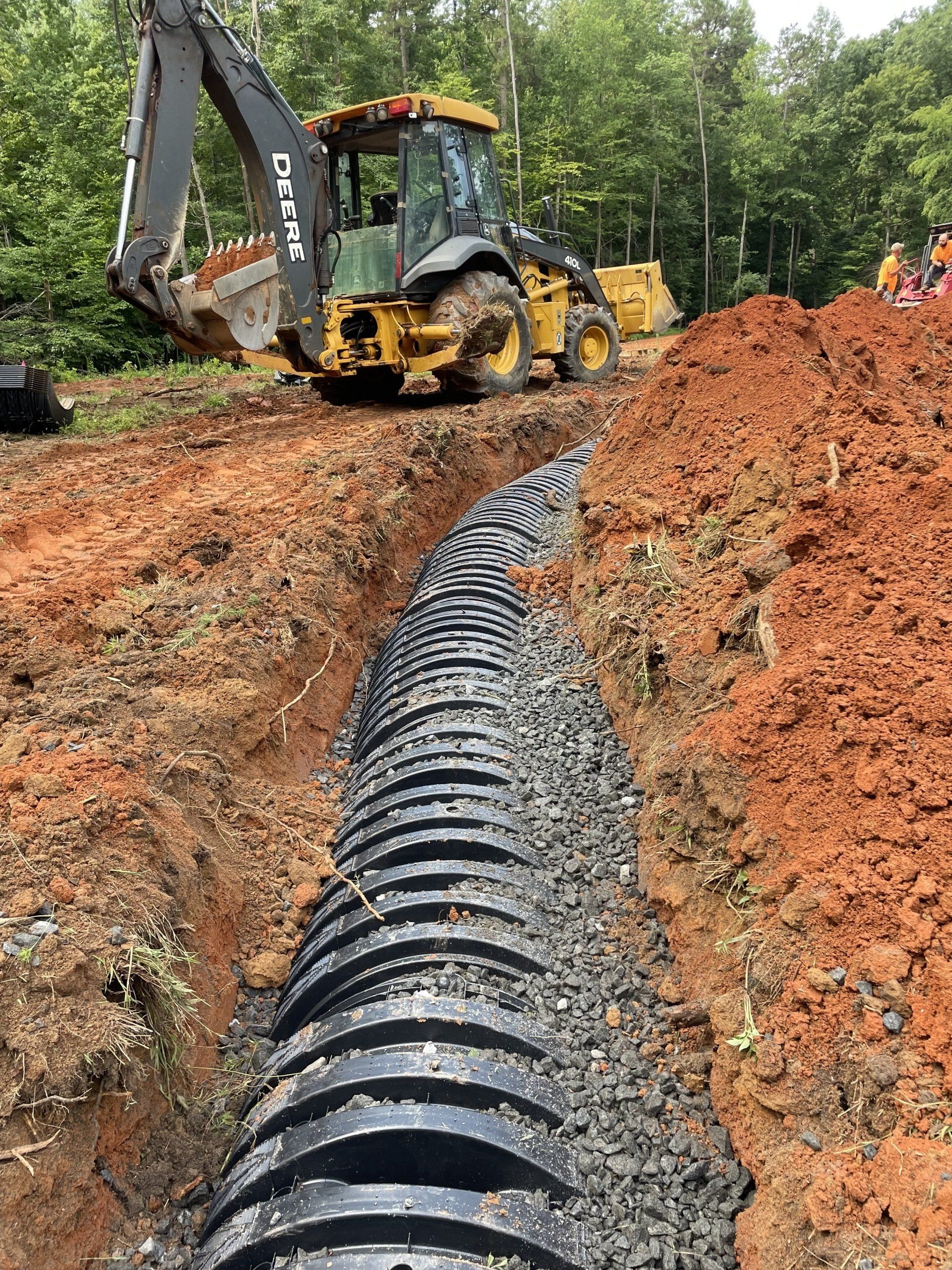 A yellow backhoe digging a trench around a black corrugated drainage pipe in a dirt field.
