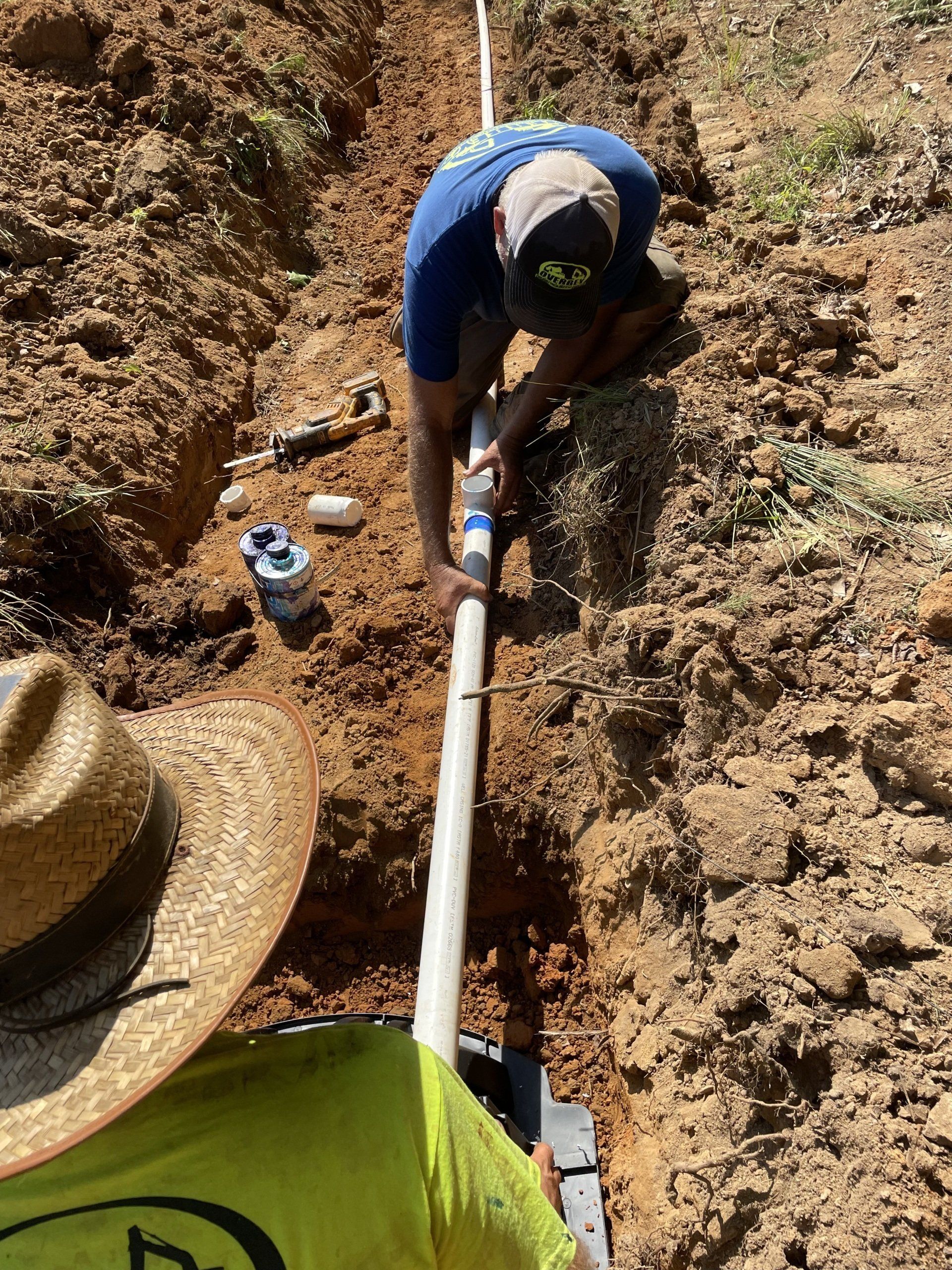Two workers installing PVC pipe in a trench; one wearing a straw hat, the other a blue shirt.