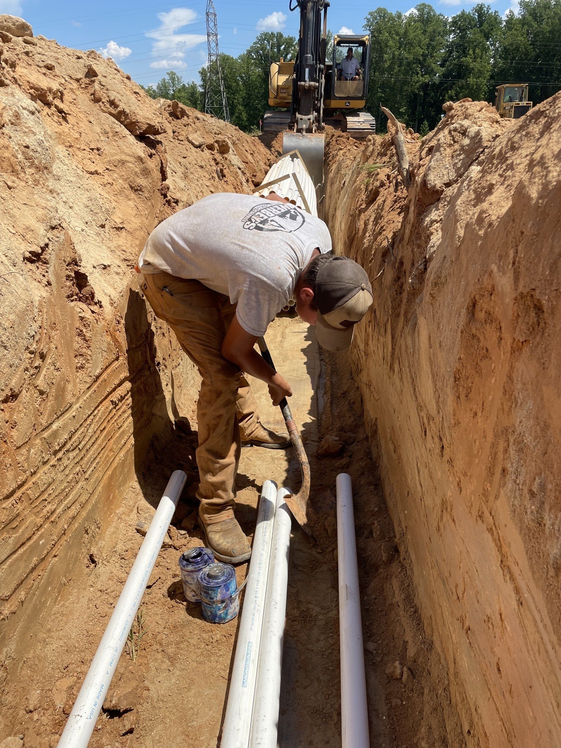 A construction worker in a trench laying pipes; an excavator is in the background.