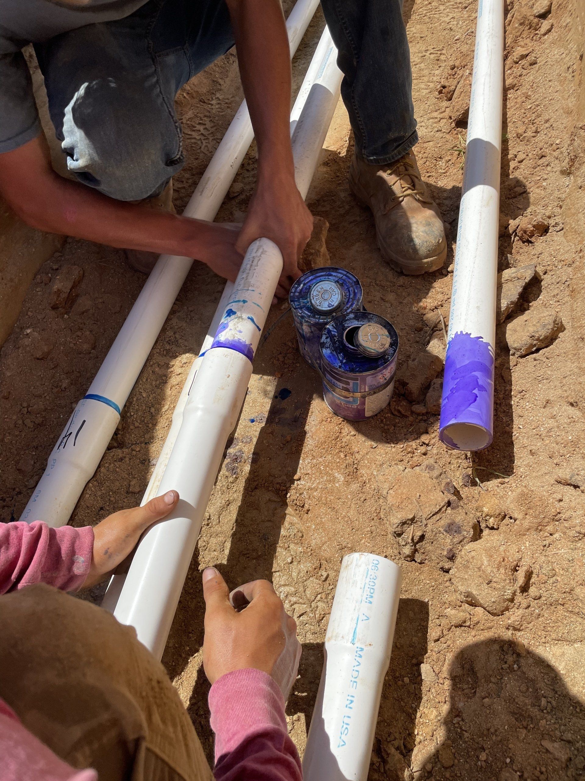 People connecting PVC pipes, using glue, in a trench.