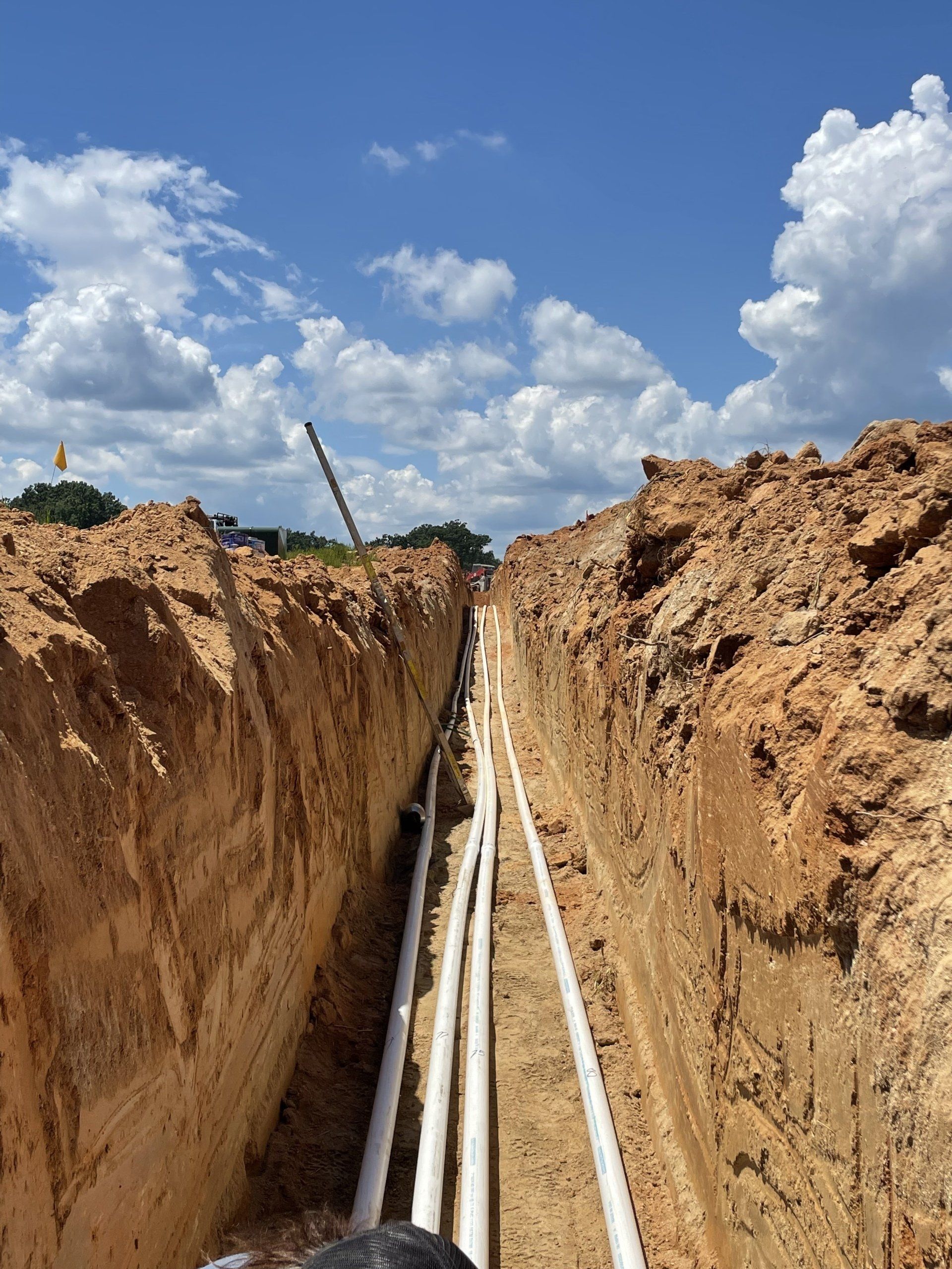 Trench with white pipes, dug in brown earth, under a blue sky with clouds; likely utility construction.