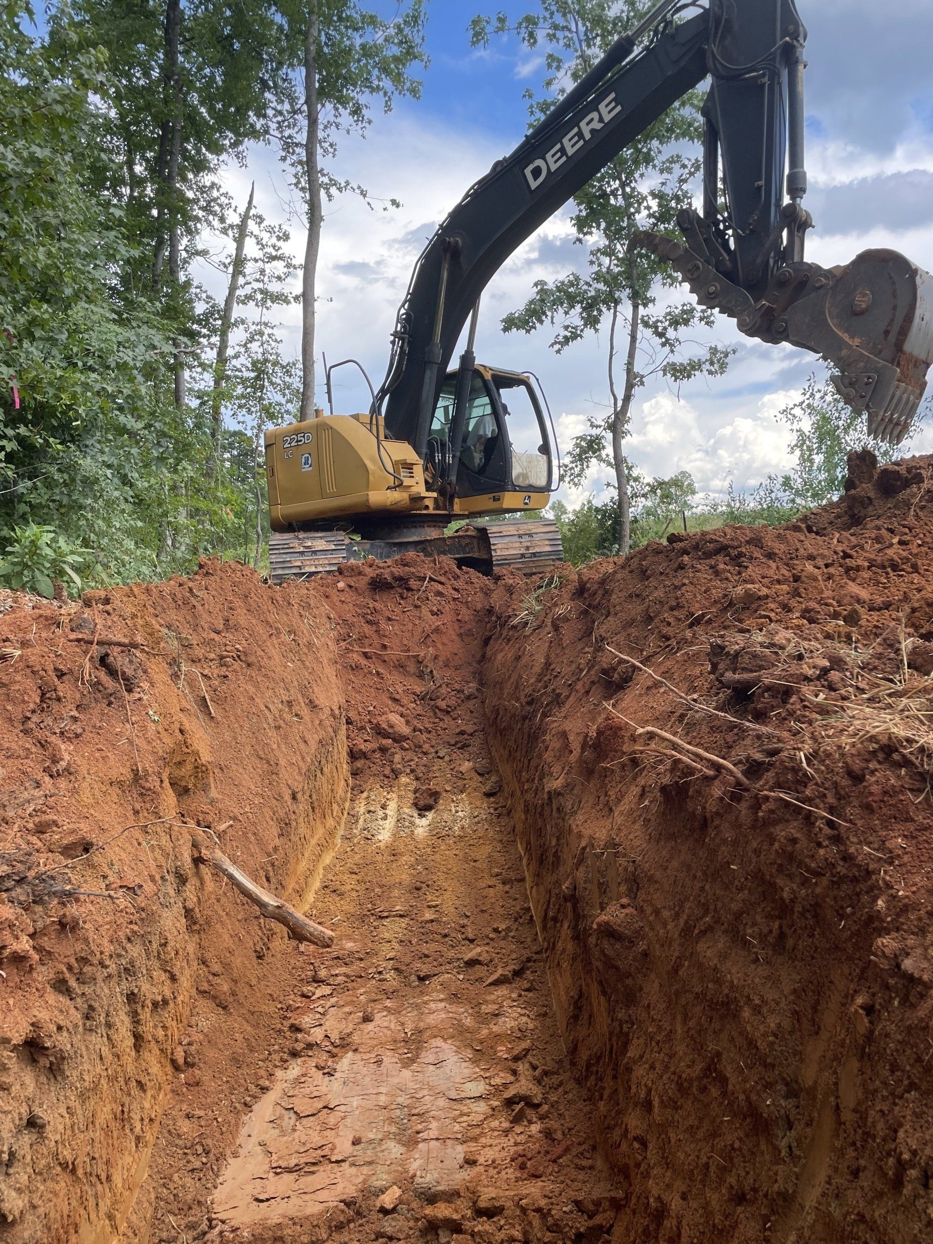 An excavator digs a trench in reddish-brown soil, with green trees and a blue sky in the background.