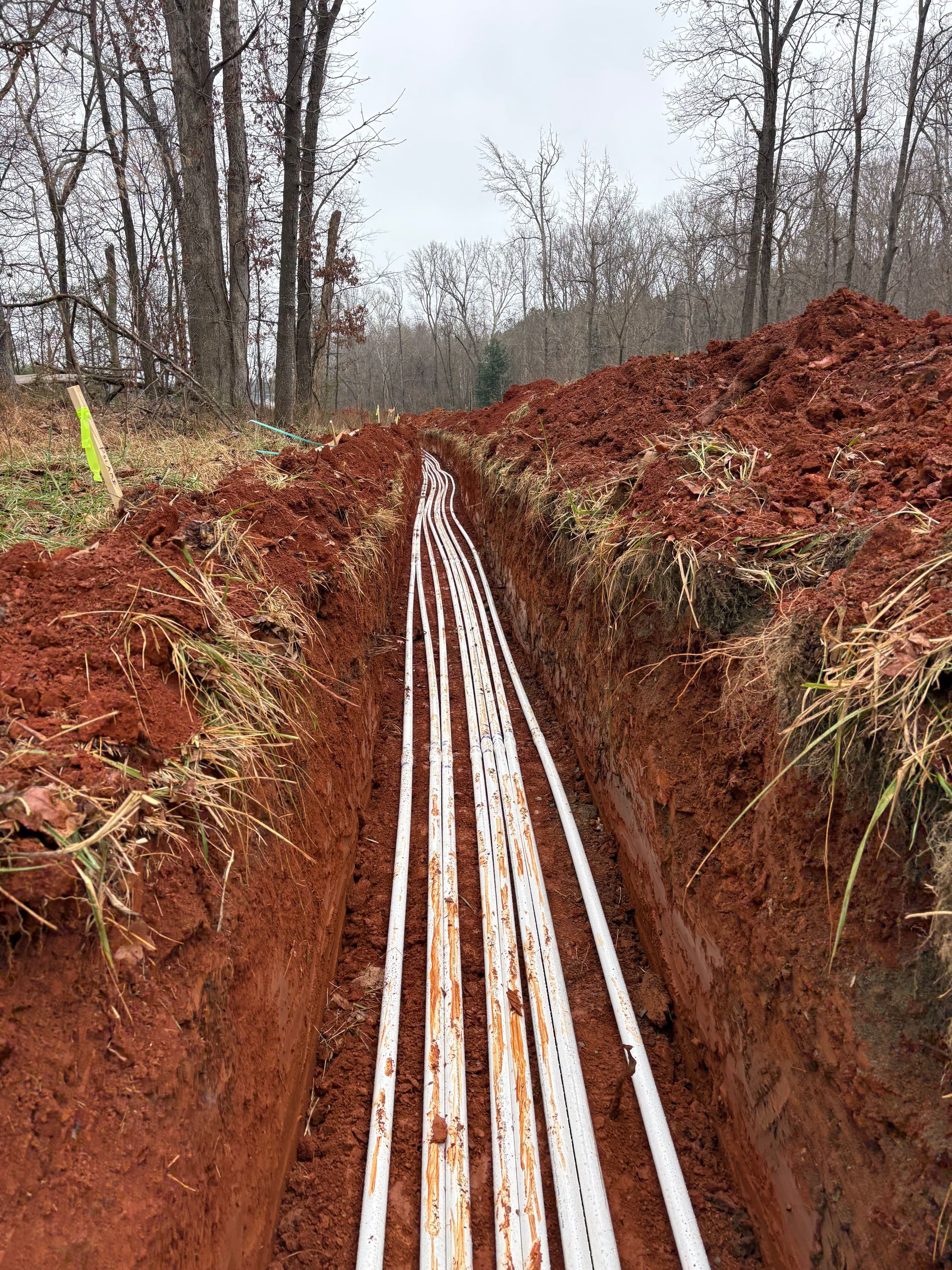 Several white plastic pipes laid in a narrow, deep red-dirt trench running through a wooded area.