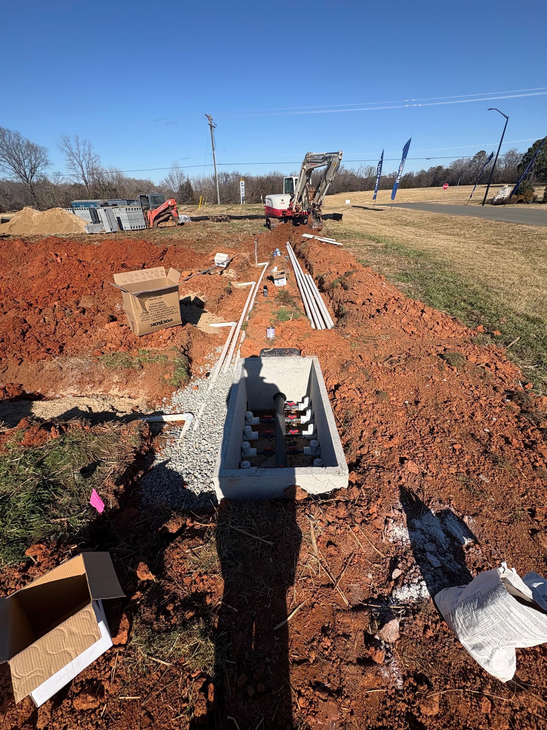 A construction site with exposed red dirt, a concrete drain basin, and construction equipment in the background.