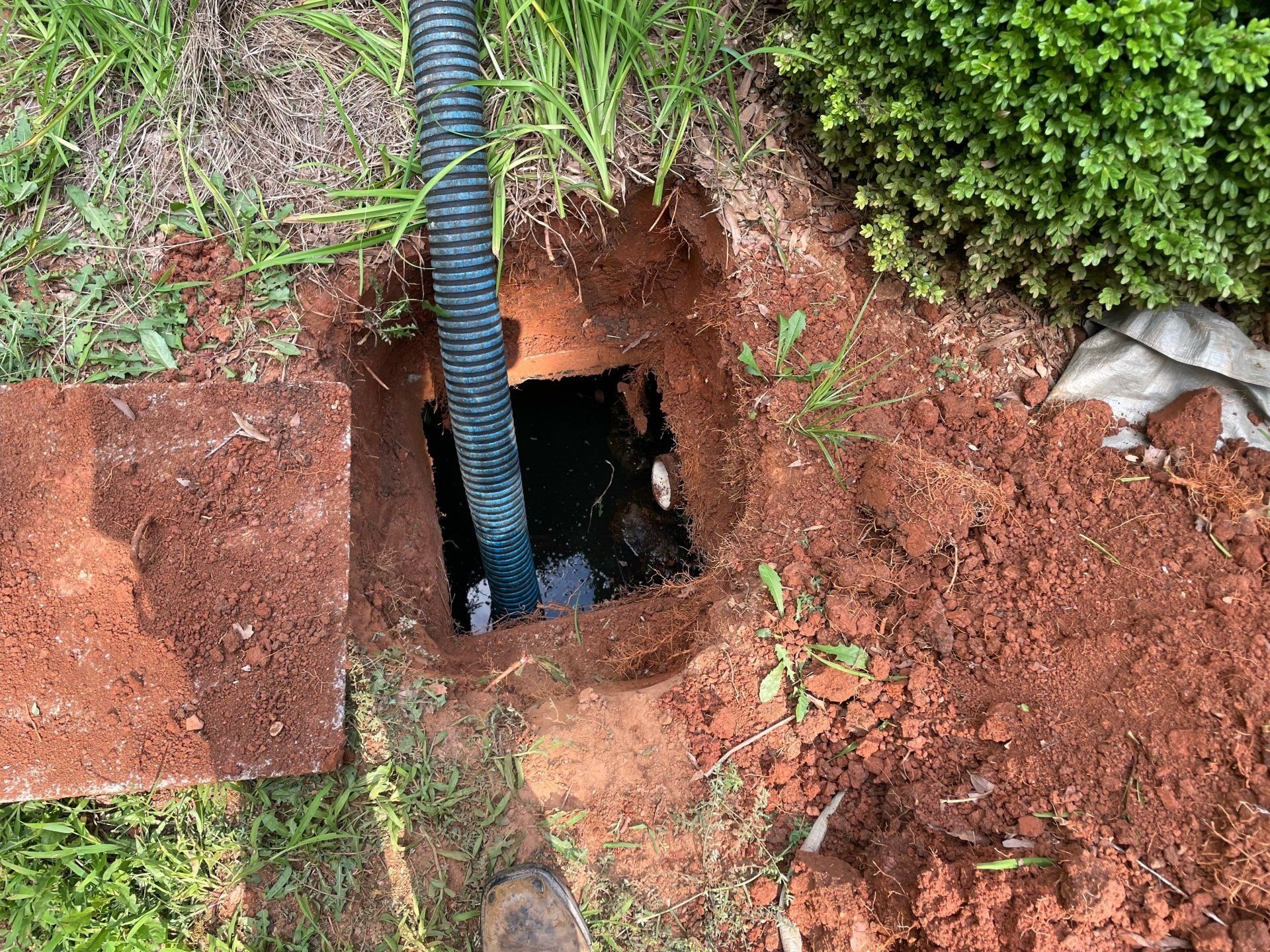 Open square hole in red clay soil with black pipe, beside brick and green bush.
