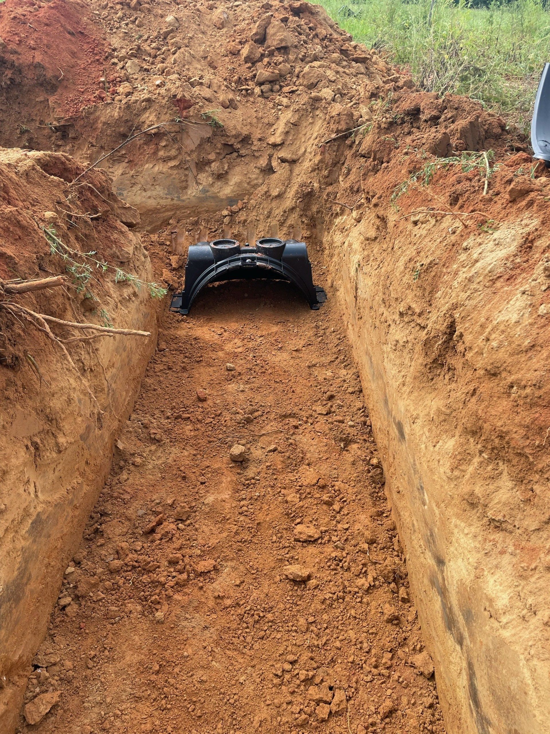 A black drainage pipe sits in a trench dug in red soil.