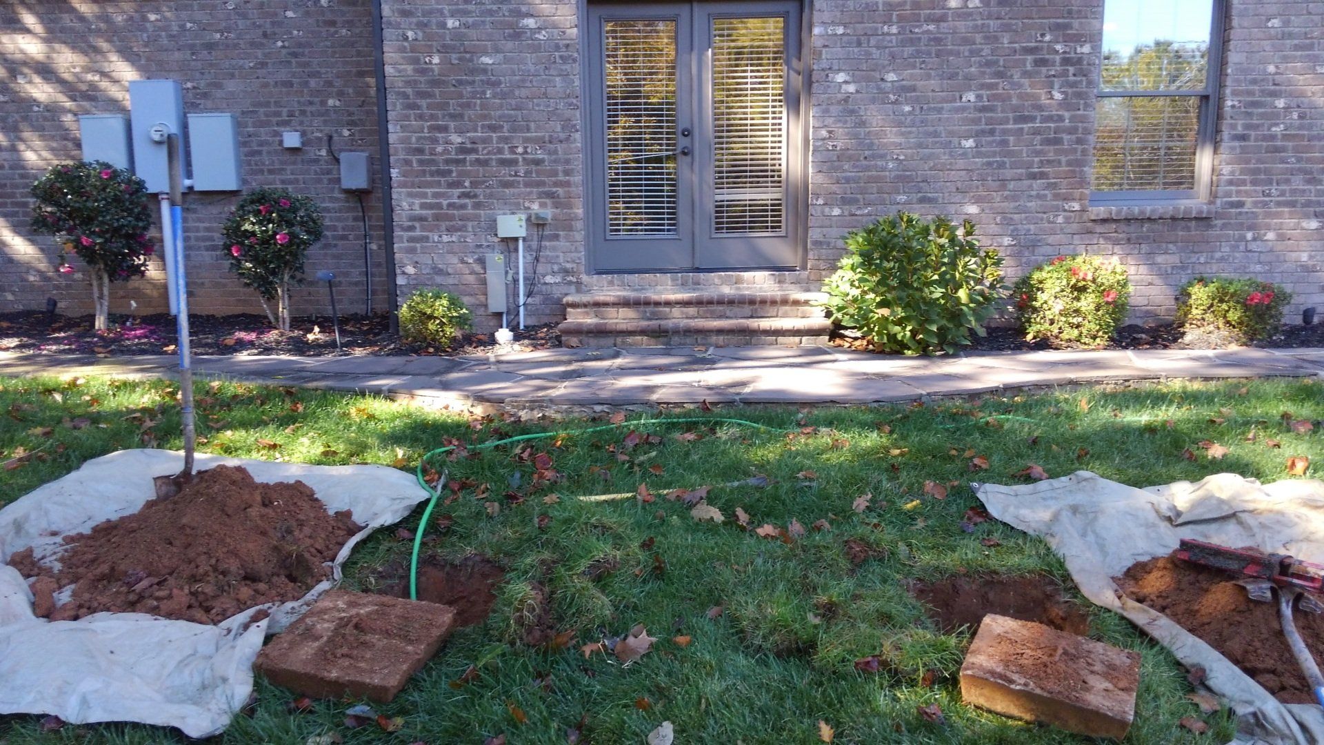 Backyard landscaping in progress with dirt piles, bricks, and a green hose. Brick house in background.