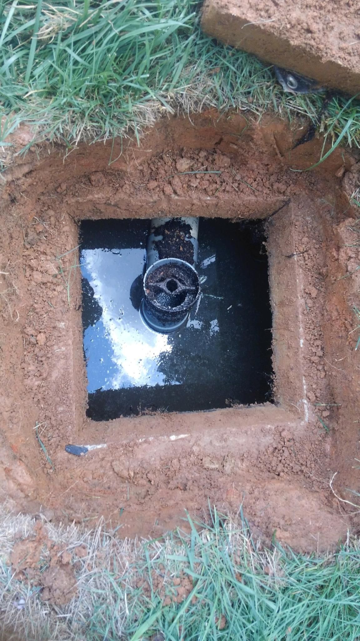 Square hole in the ground with water and pipe fitting surrounded by dirt and grass.
