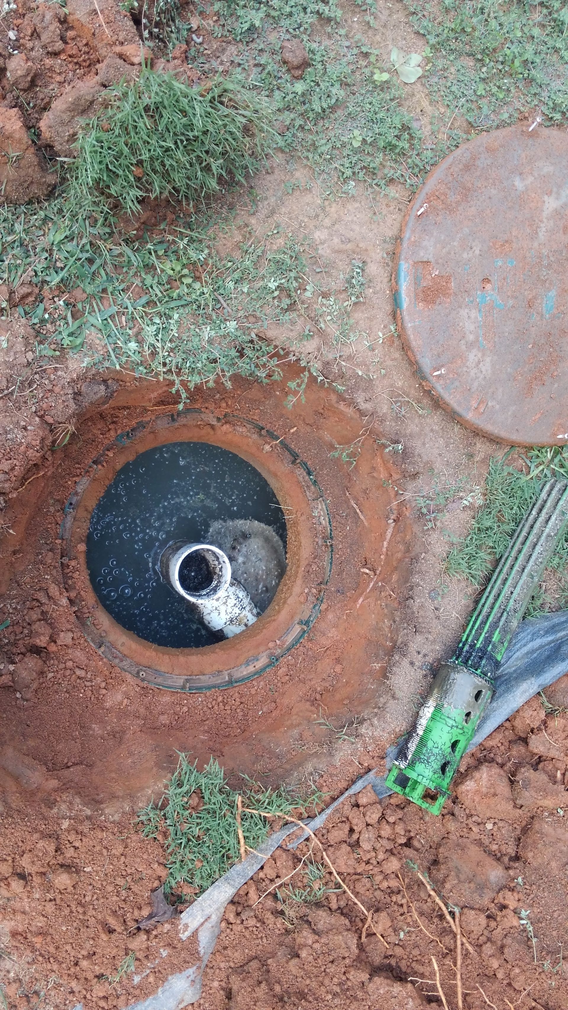 Open well with metal cover removed, surrounded by dirt and grass. Sprinkler nozzle in frame.