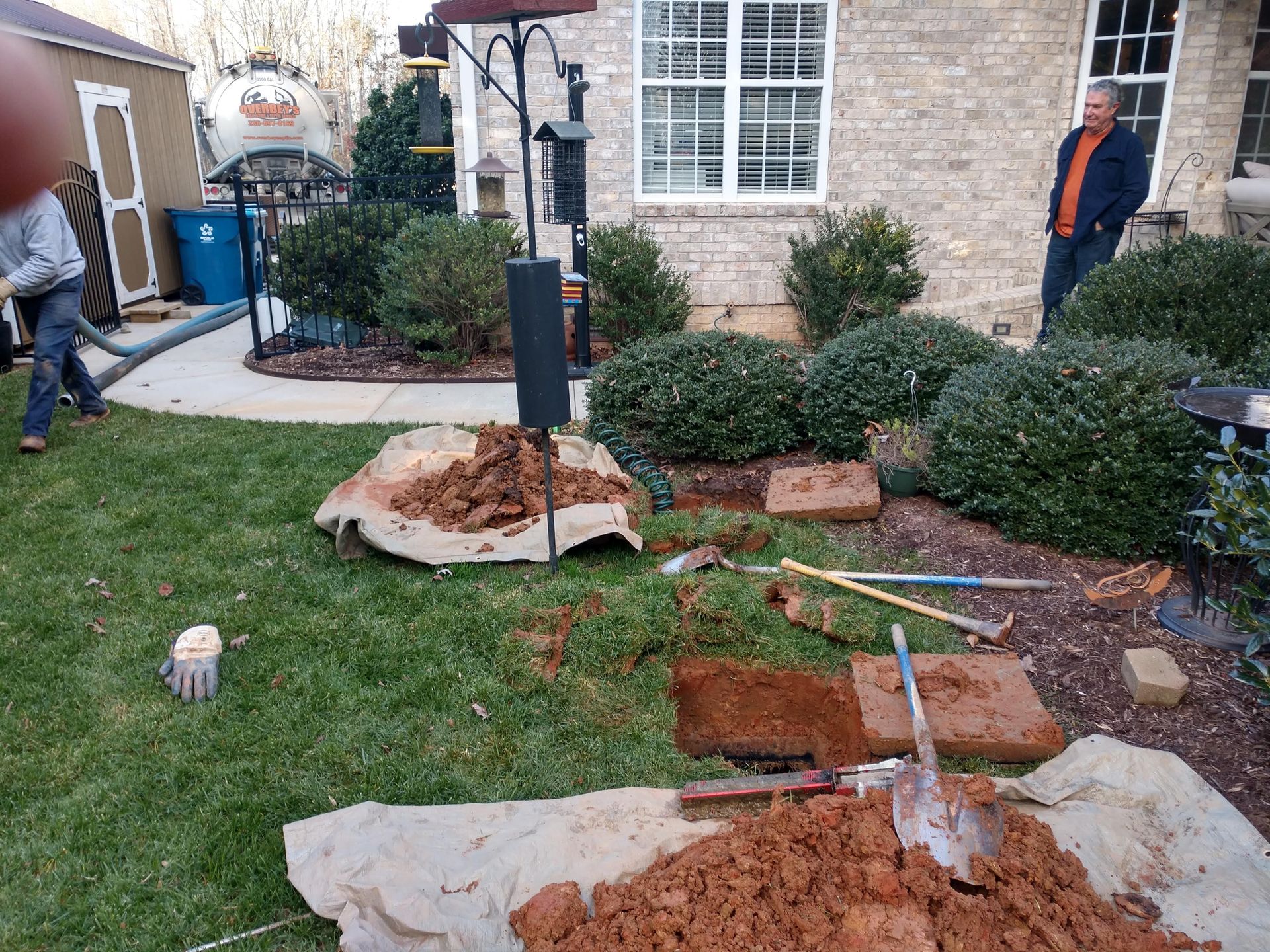 Yard work in progress: Dirt pile, exposed ground, bricks, bushes, and two people near a house.