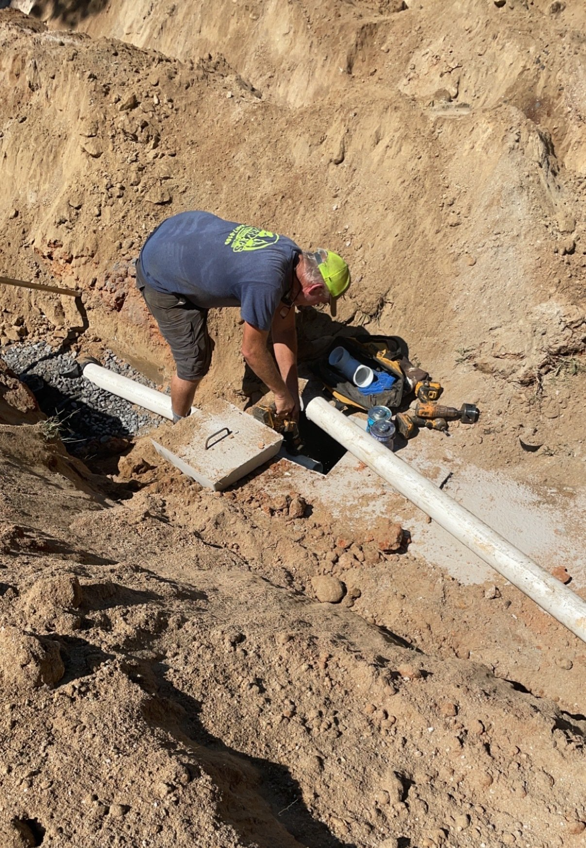 Man installing pipes in an excavated trench. He wears a blue shirt and green cap, surrounded by dirt and gravel.