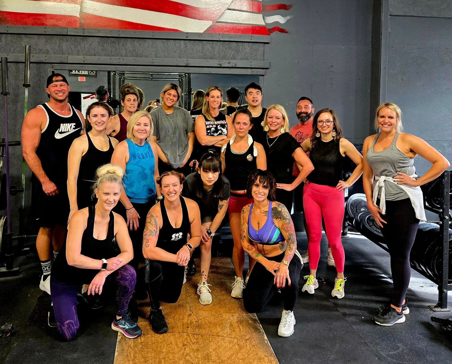 Group of people in gym; posing, smiling. Weights, flag, mirror.