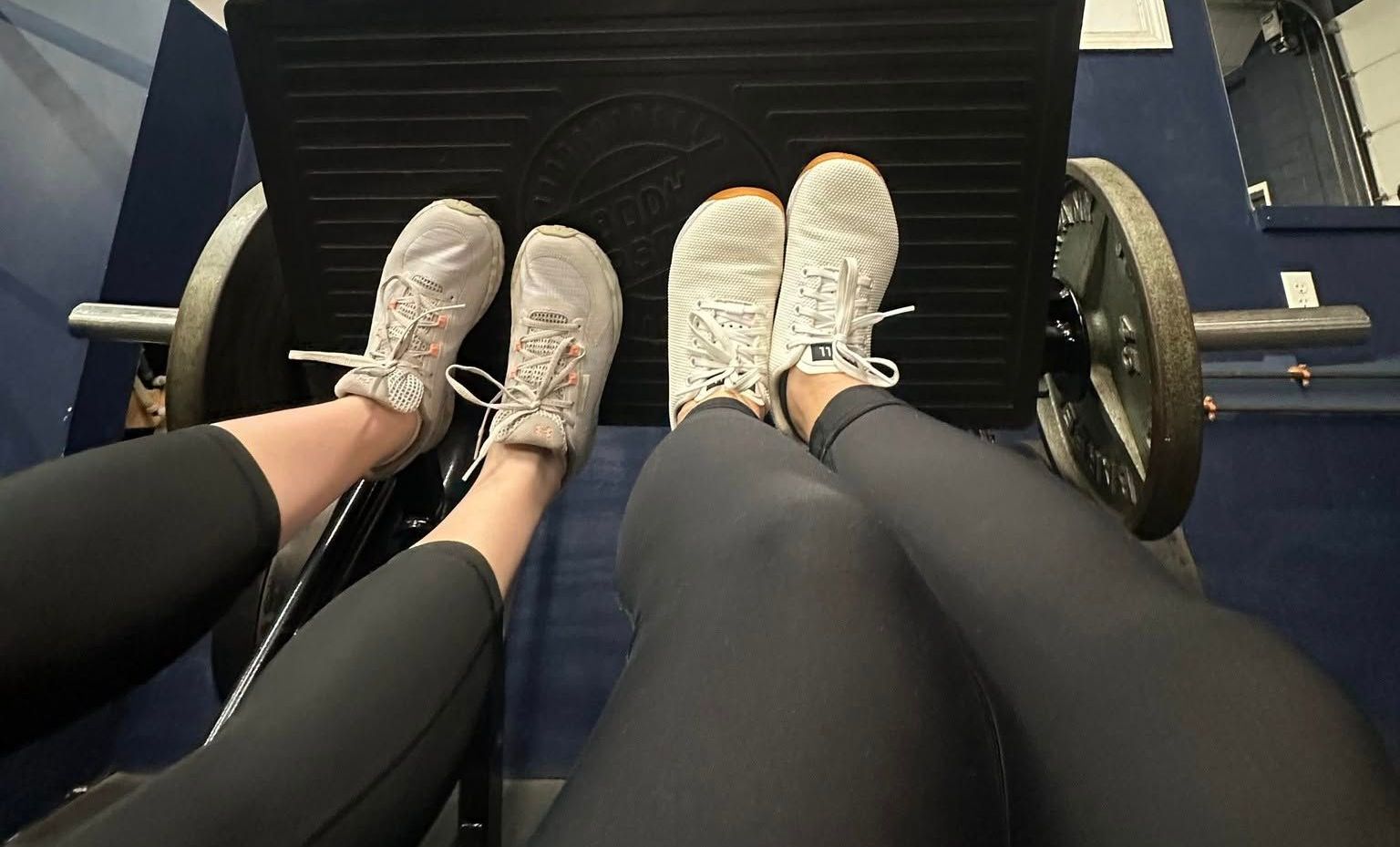 Two people's legs and feet in sneakers on a leg press machine at a gym, working out.
