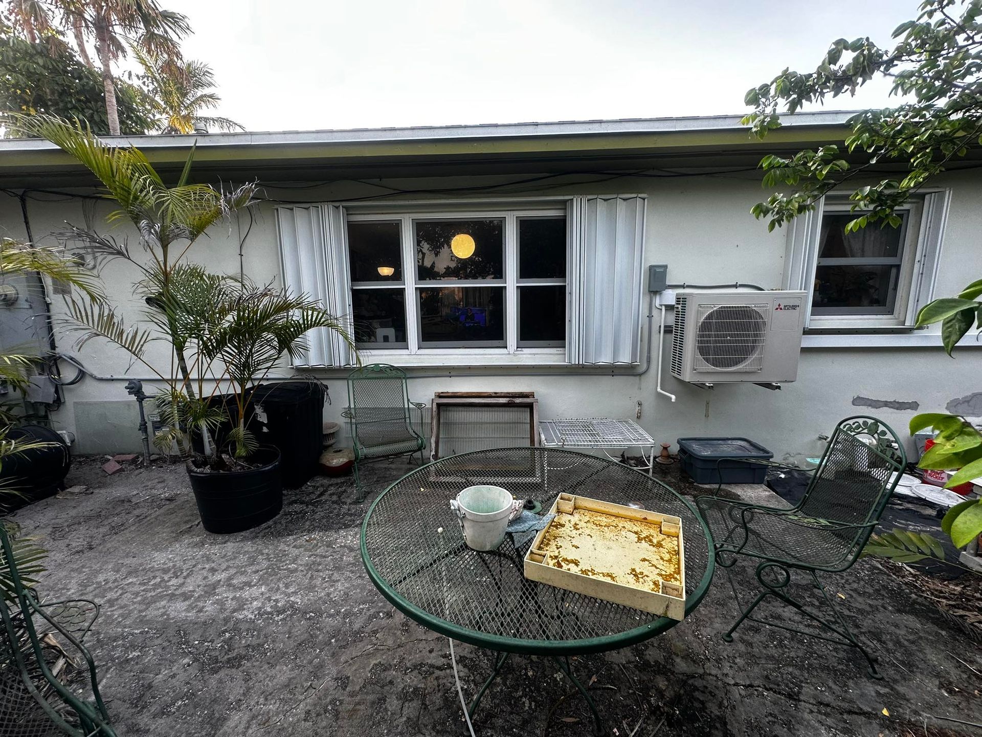 Back patio with table, chairs, fan, and window of a white building; palm trees visible.