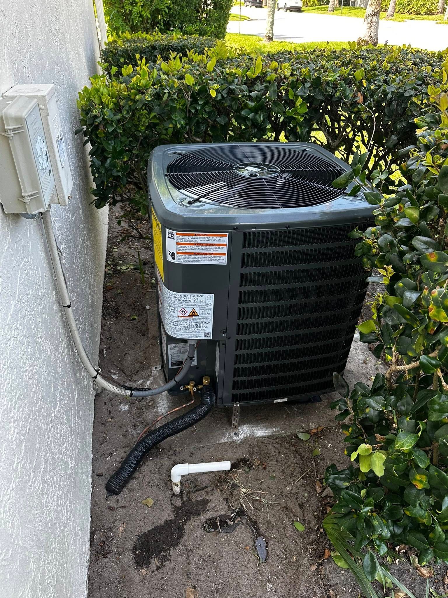 Outdoor AC unit next to a white wall and shrubbery. Hoses and a white pipe are visible on the ground.