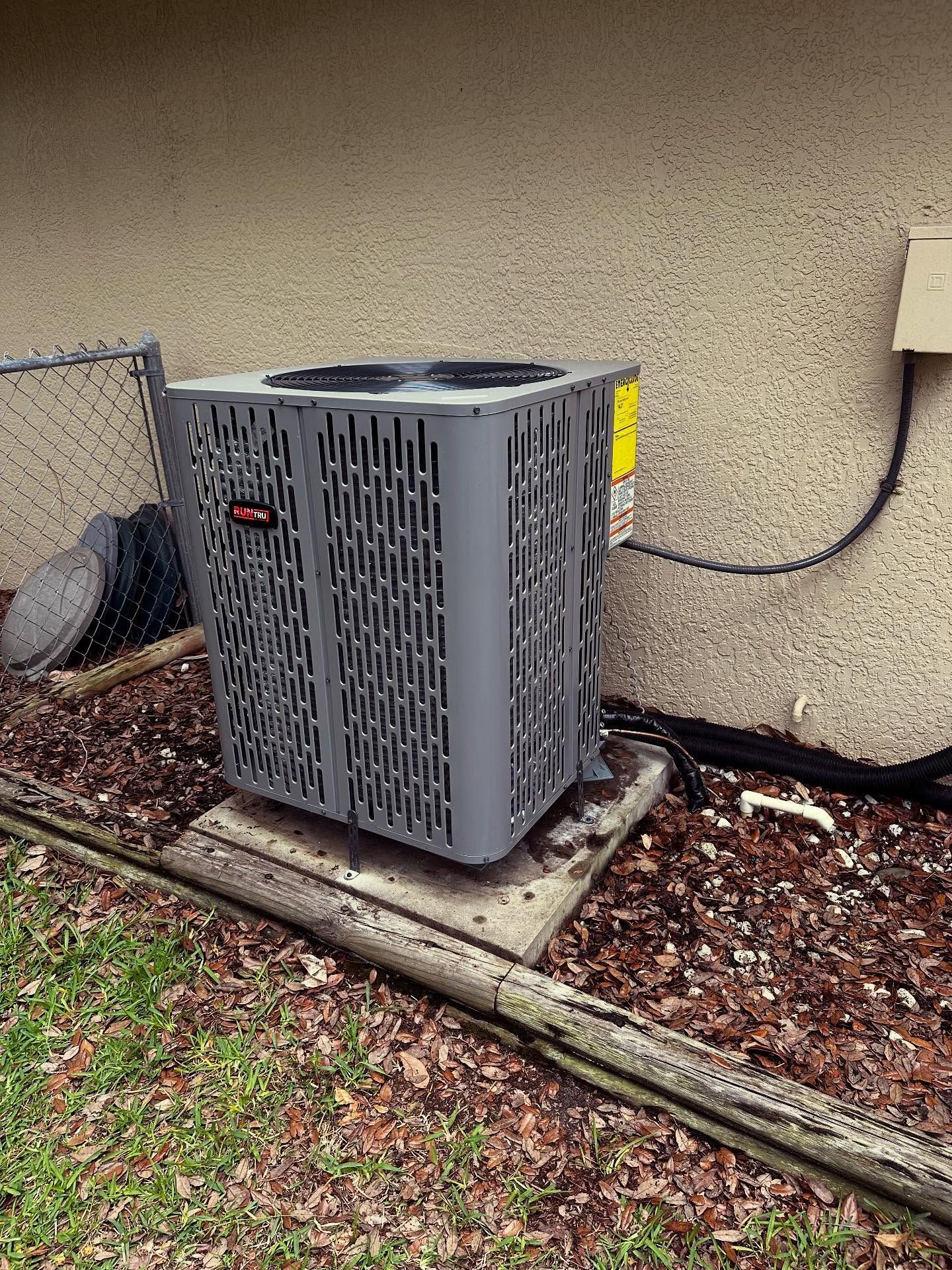 Gray air conditioning unit outside a building, set on a concrete pad with surrounding mulch and a wooden border.