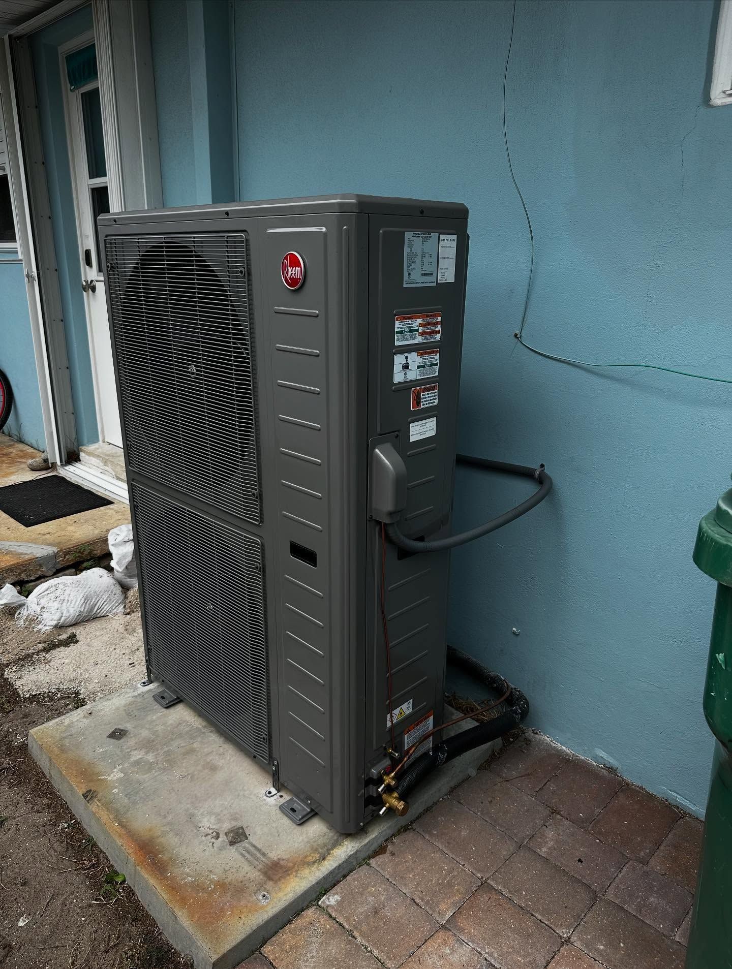 A gray Rheem heat pump unit outside a blue building, on a concrete pad.