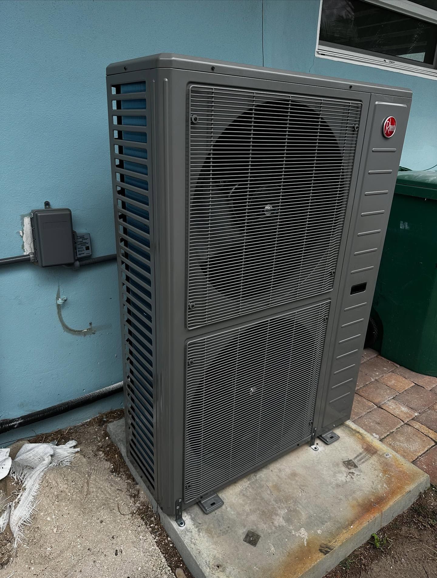 Dark gray Rheem air conditioning unit outside a blue building, next to a green trash can.
