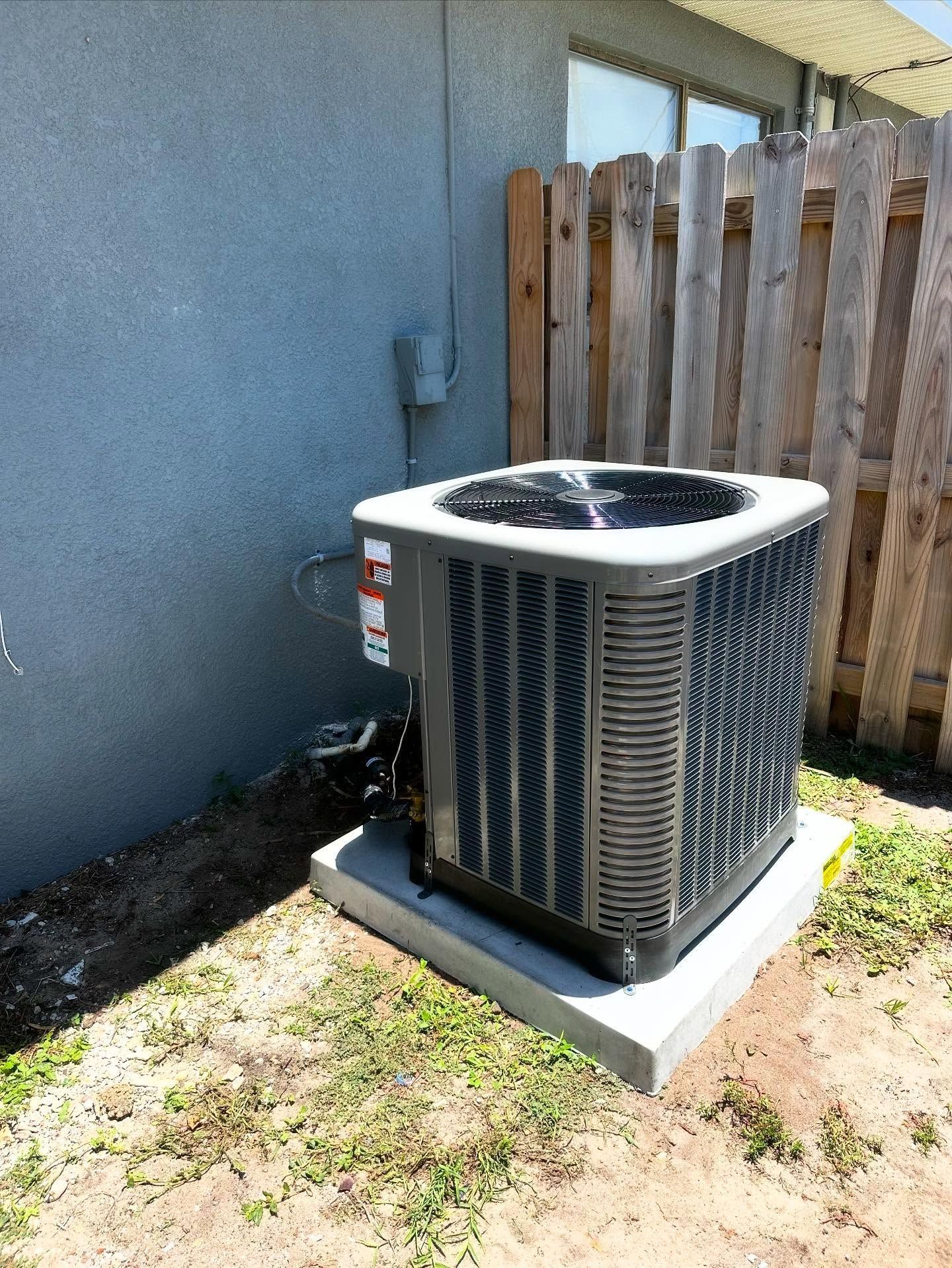 Air conditioning unit on a concrete pad next to a gray wall and wooden fence.