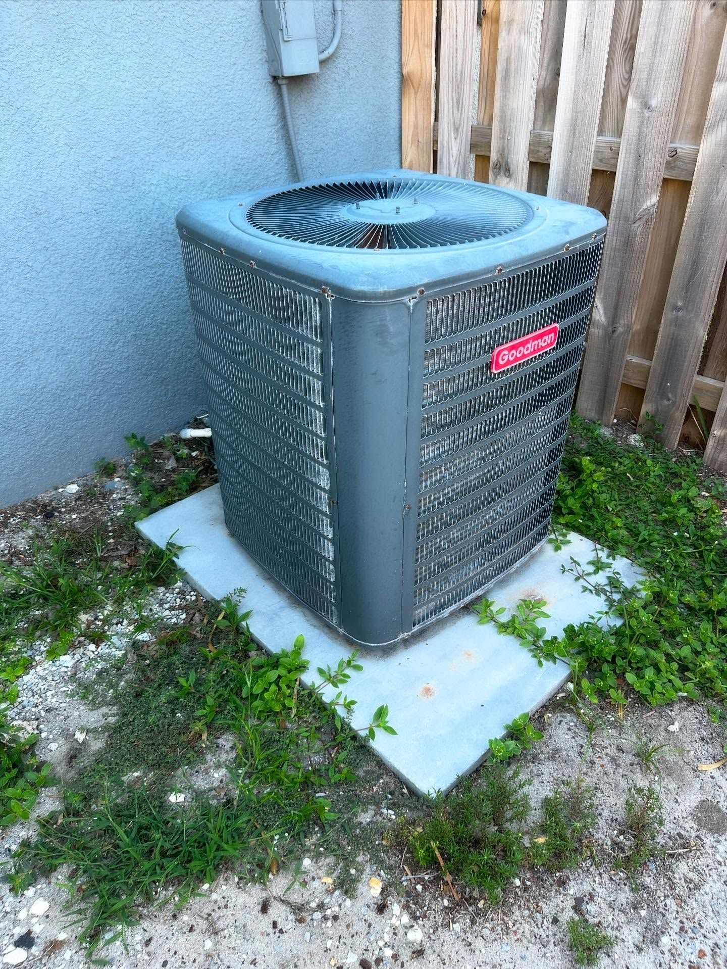 Air conditioning unit on a concrete pad near a wall and wooden fence, surrounded by weeds.