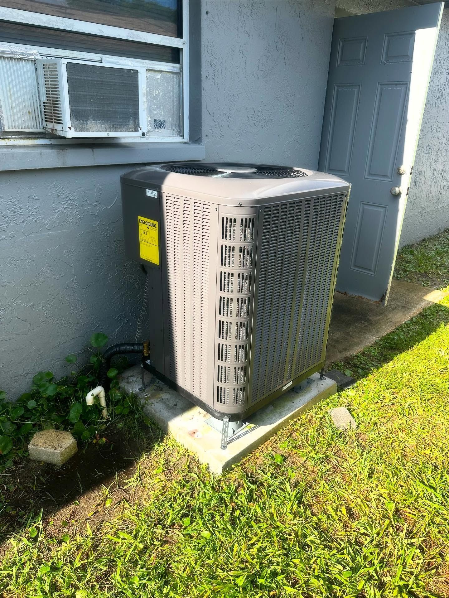 HVAC unit outside a building with an air conditioner and open door; green grass surrounds.