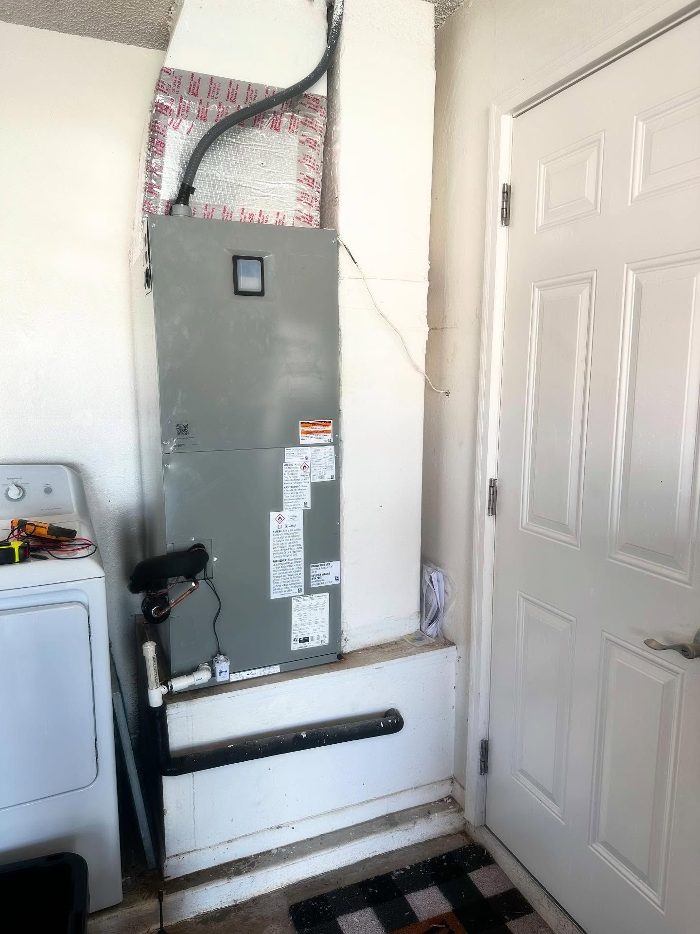 HVAC unit next to a white door and a washing machine in a room with white walls and a dark mat.