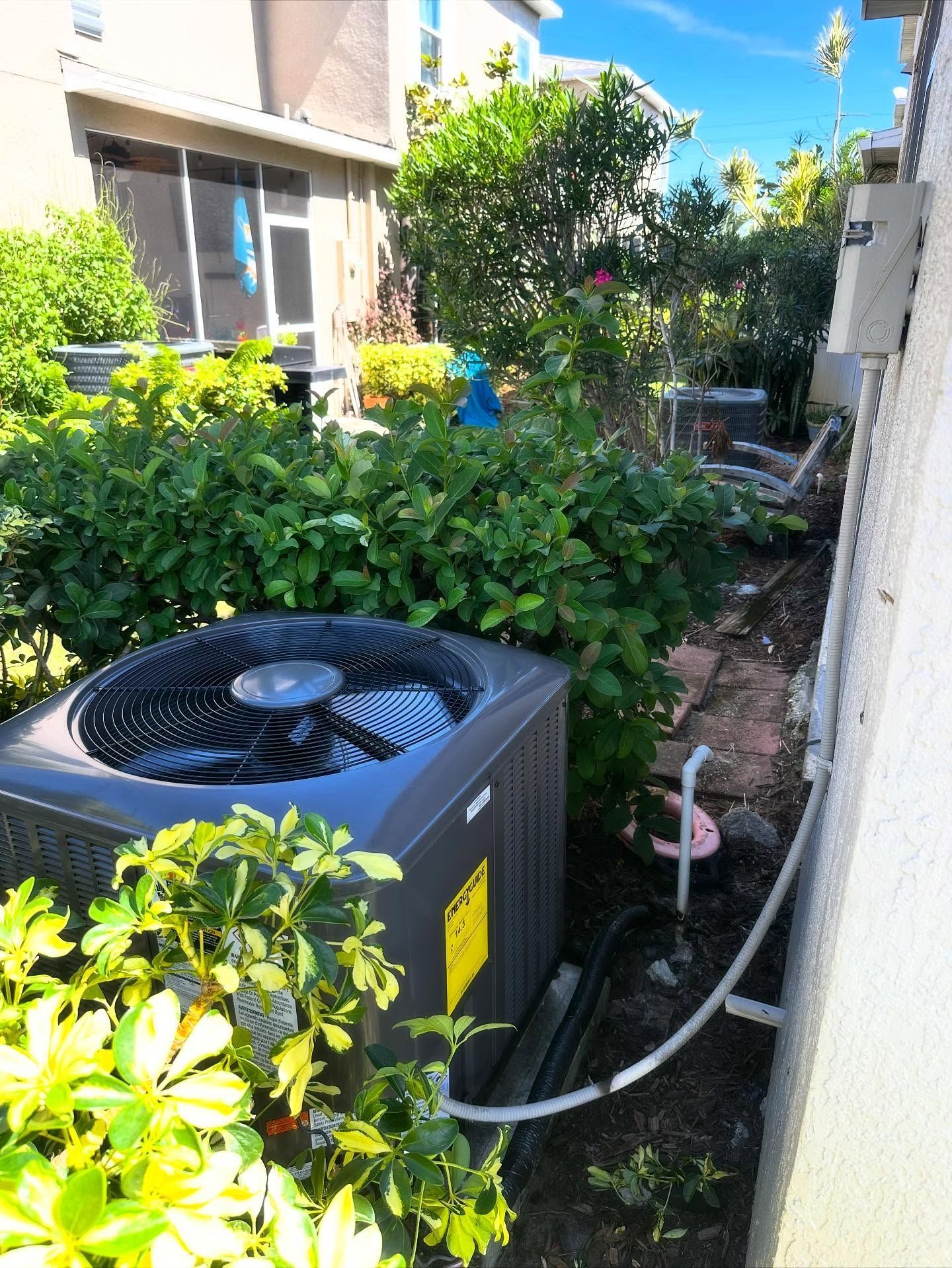Air conditioning unit surrounded by green bushes and plants next to a building.