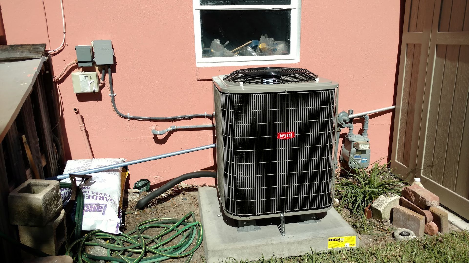 An air conditioning unit next to a building wall with electrical components and a window.