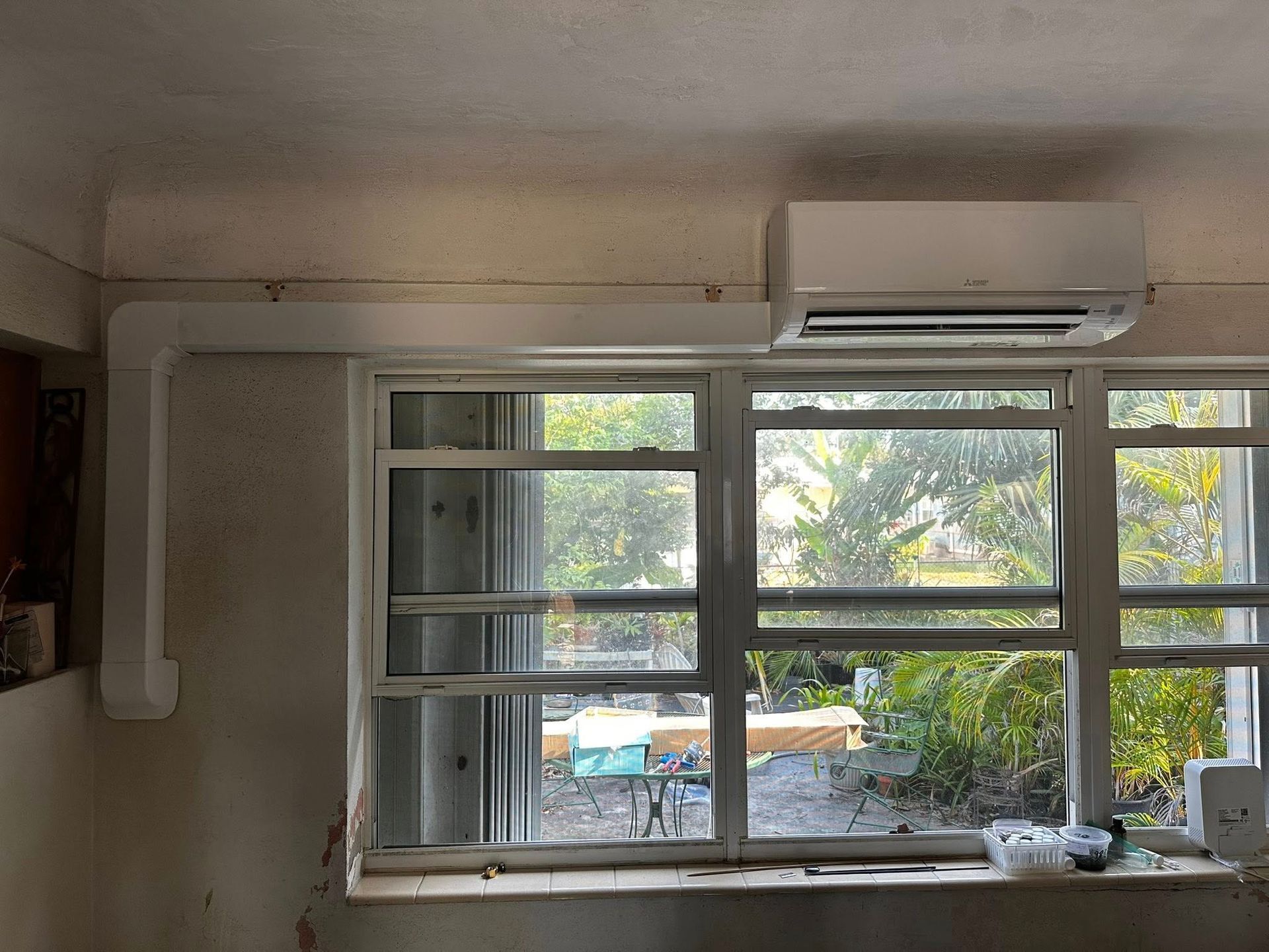Air conditioner mounted above a window, with white ducting running along the wall.