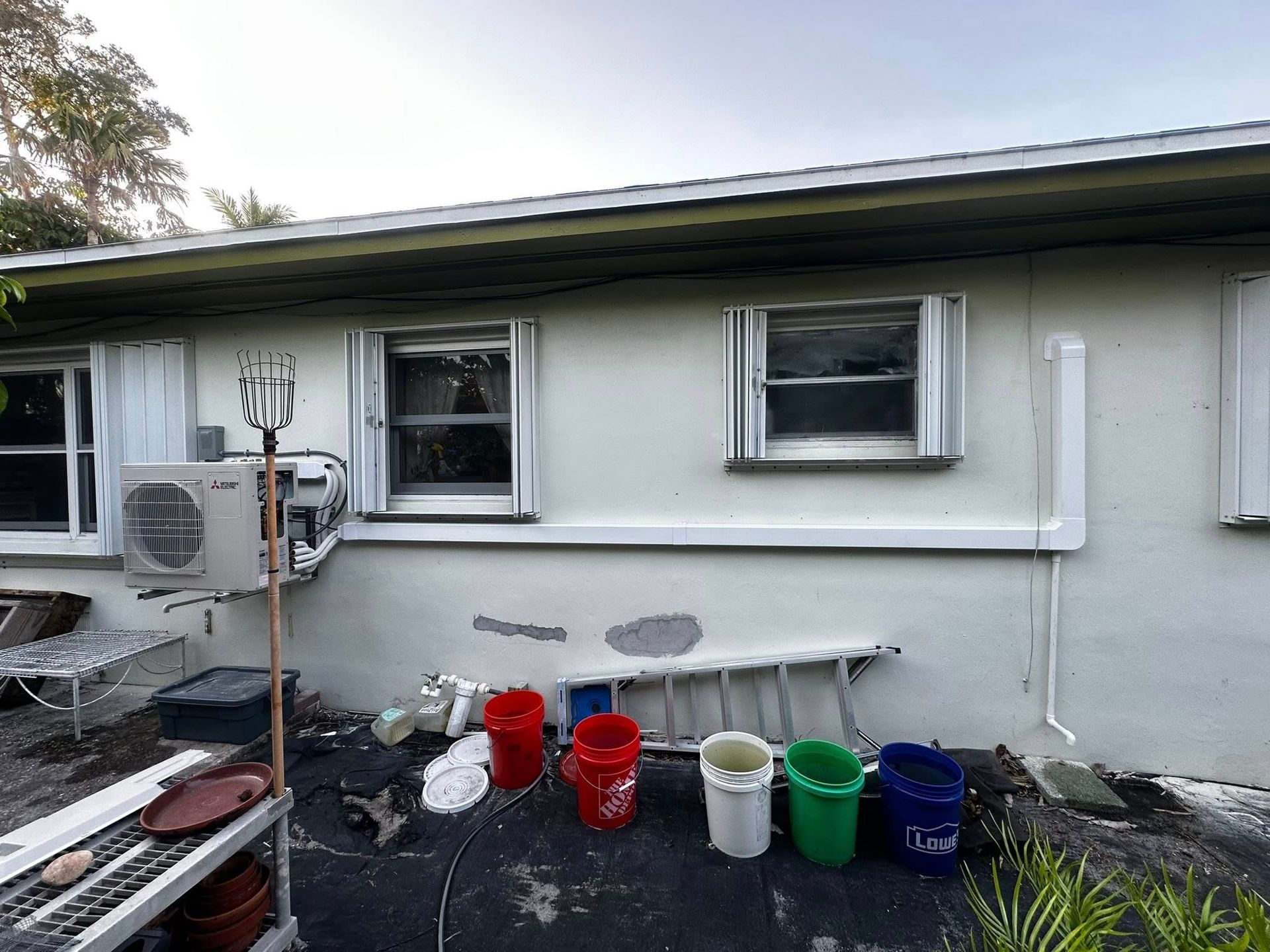 White building exterior with windows, an air conditioner, and several buckets, a ladder, and yard debris below.