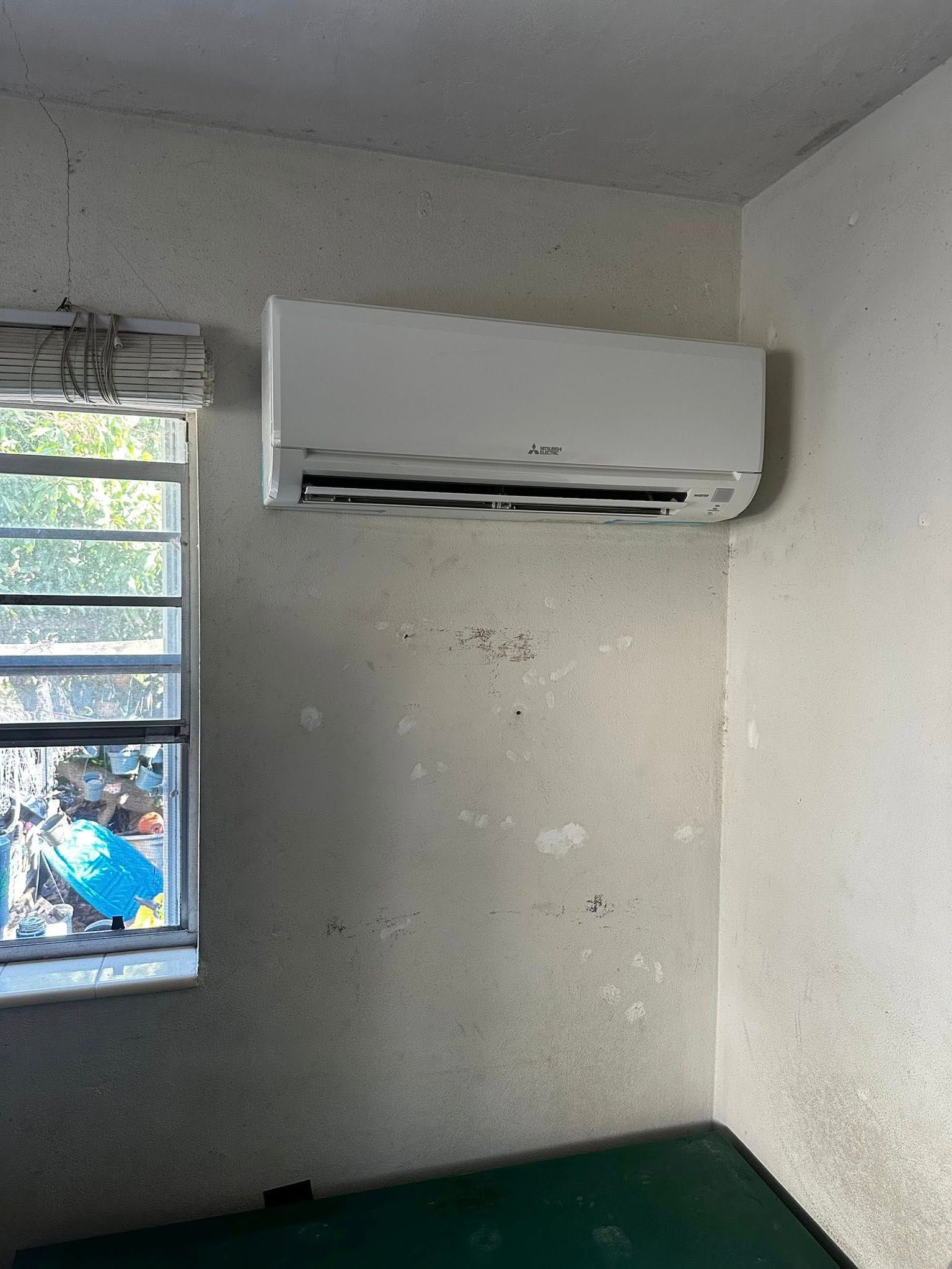 White Mitsubishi AC unit mounted on a stained wall above a green surface, near a window.