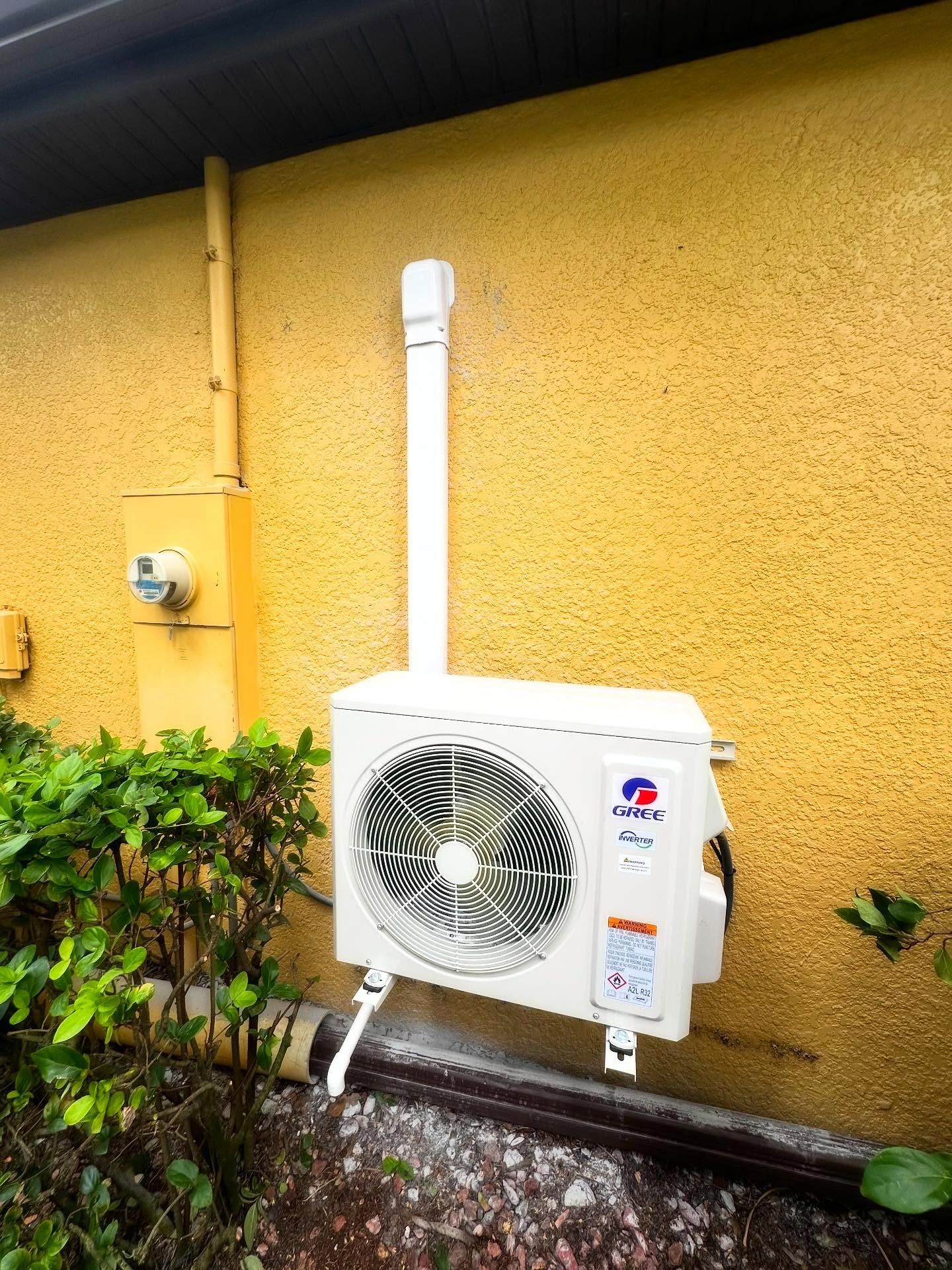 White air conditioning unit on yellow stucco wall next to greenery and a yellow electrical box.