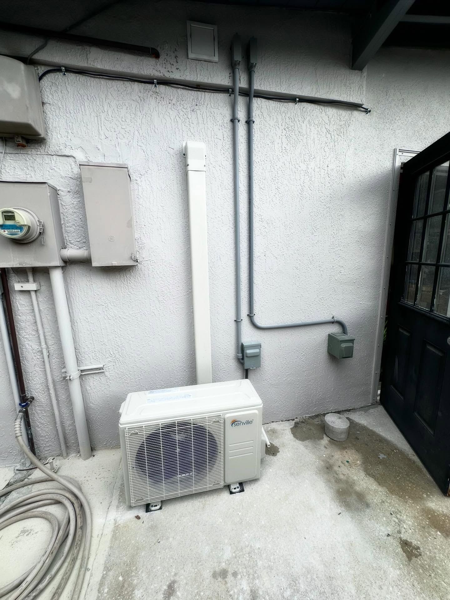 An outdoor air conditioning unit next to a white stucco wall with electrical conduits and a door.