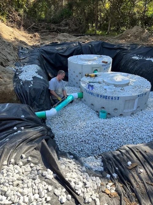 Man working on a septic system installation. Two concrete tanks surrounded by gravel and black liner in an excavated pit.