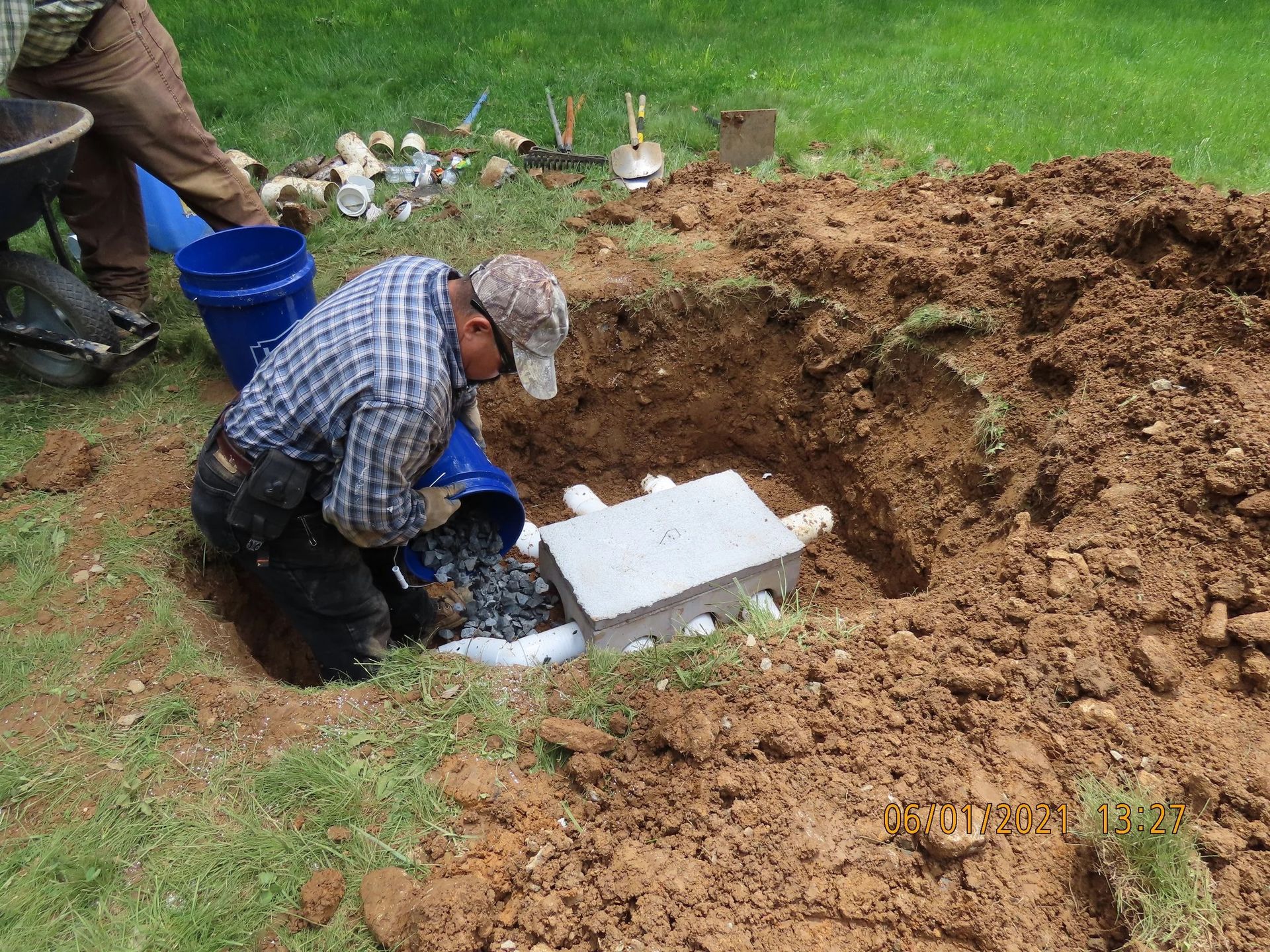 Man installing a septic tank in a dirt hole outdoors. He pours gravel from a blue bucket.