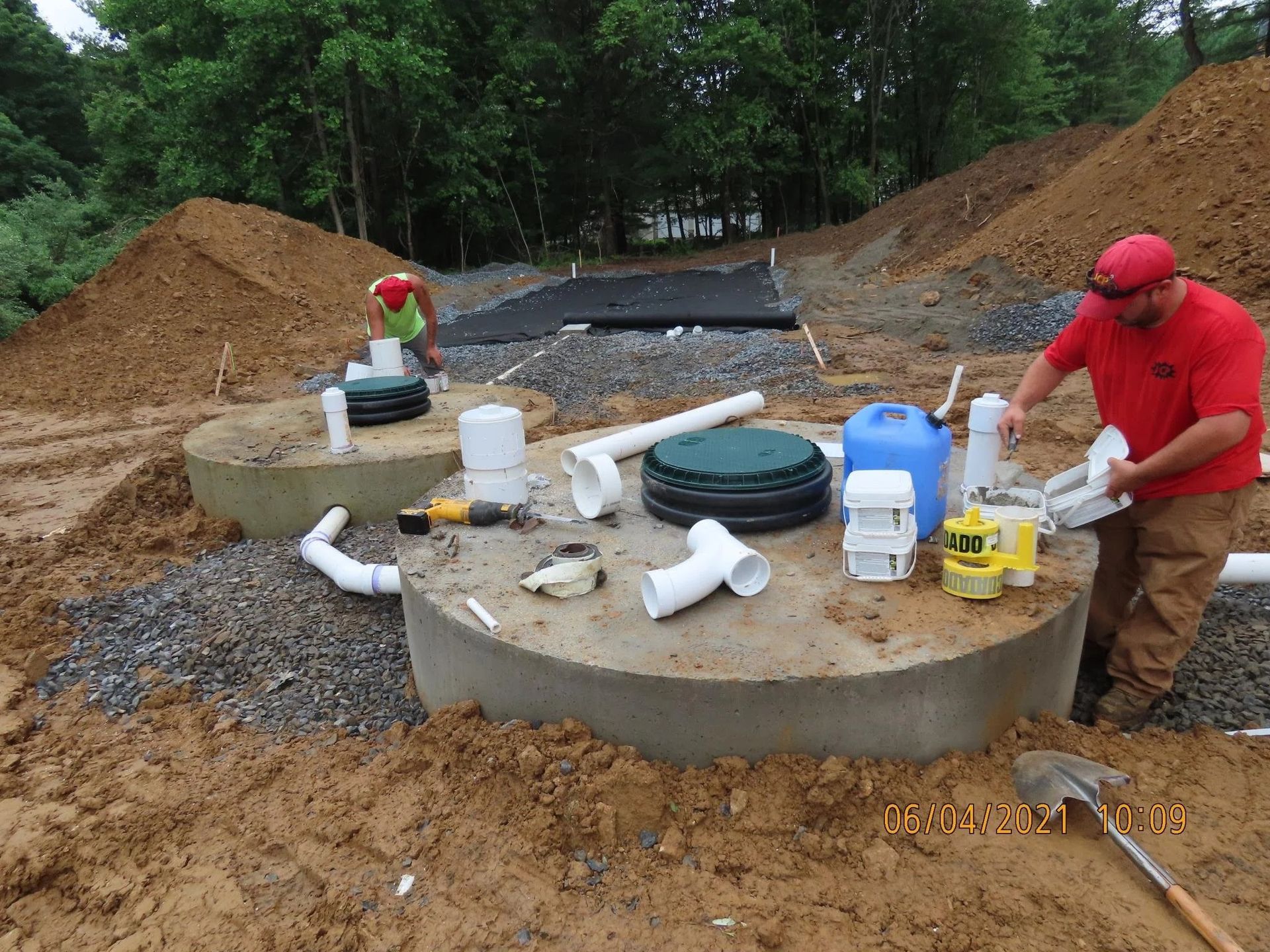 Two workers install a septic system. Gray concrete tanks are surrounded by dirt and gravel.