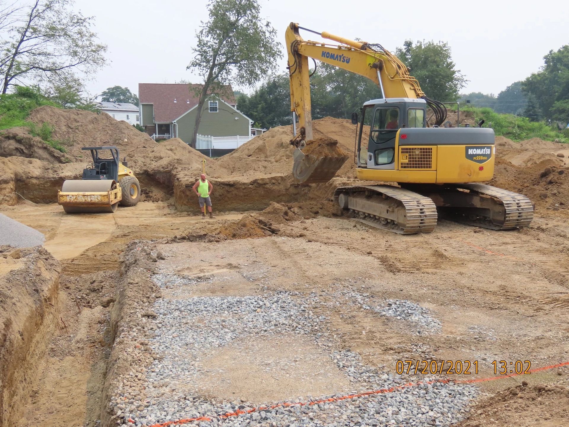 Construction site with yellow excavator digging, compact roller, man, and foundation filled with gravel.