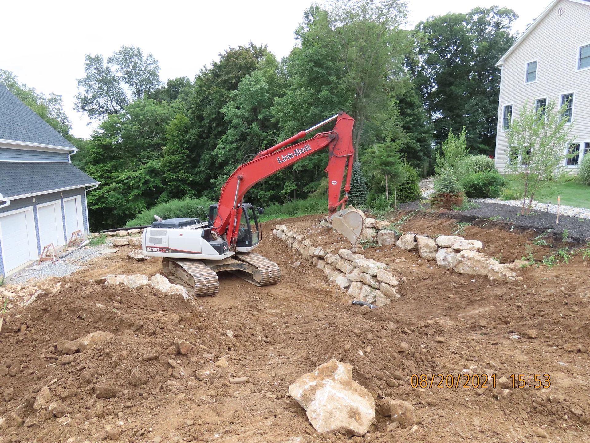 An excavator works in a yard. Red arm, white body, brown dirt, stone retaining walls, trees and a house.