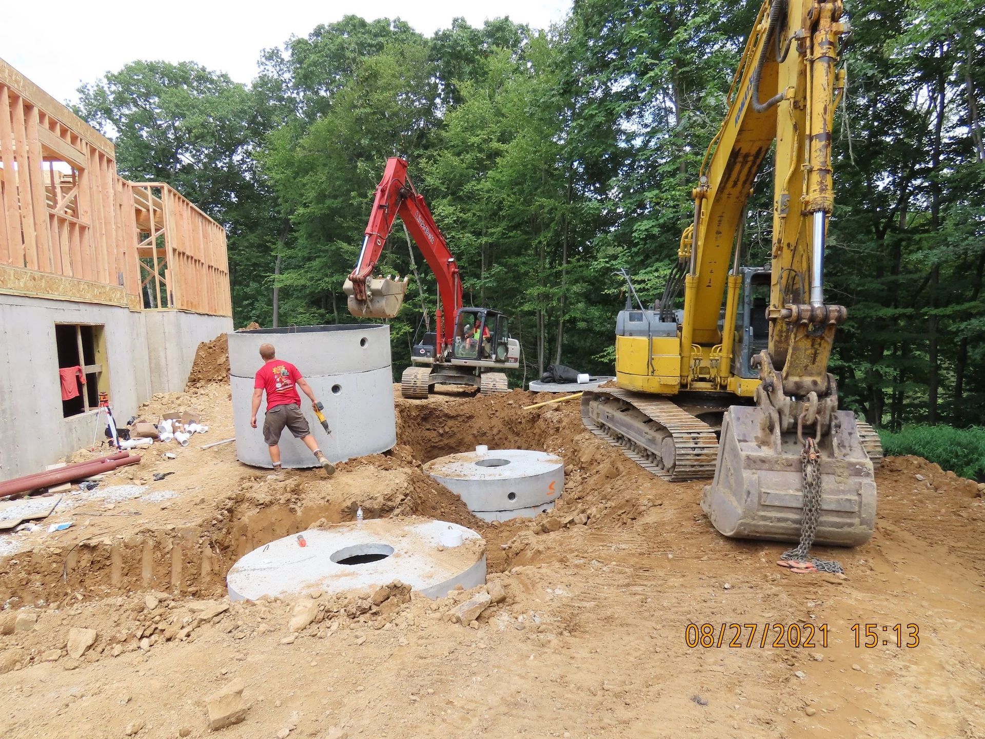 Construction site: excavators placing concrete rings, worker, unfinished house.