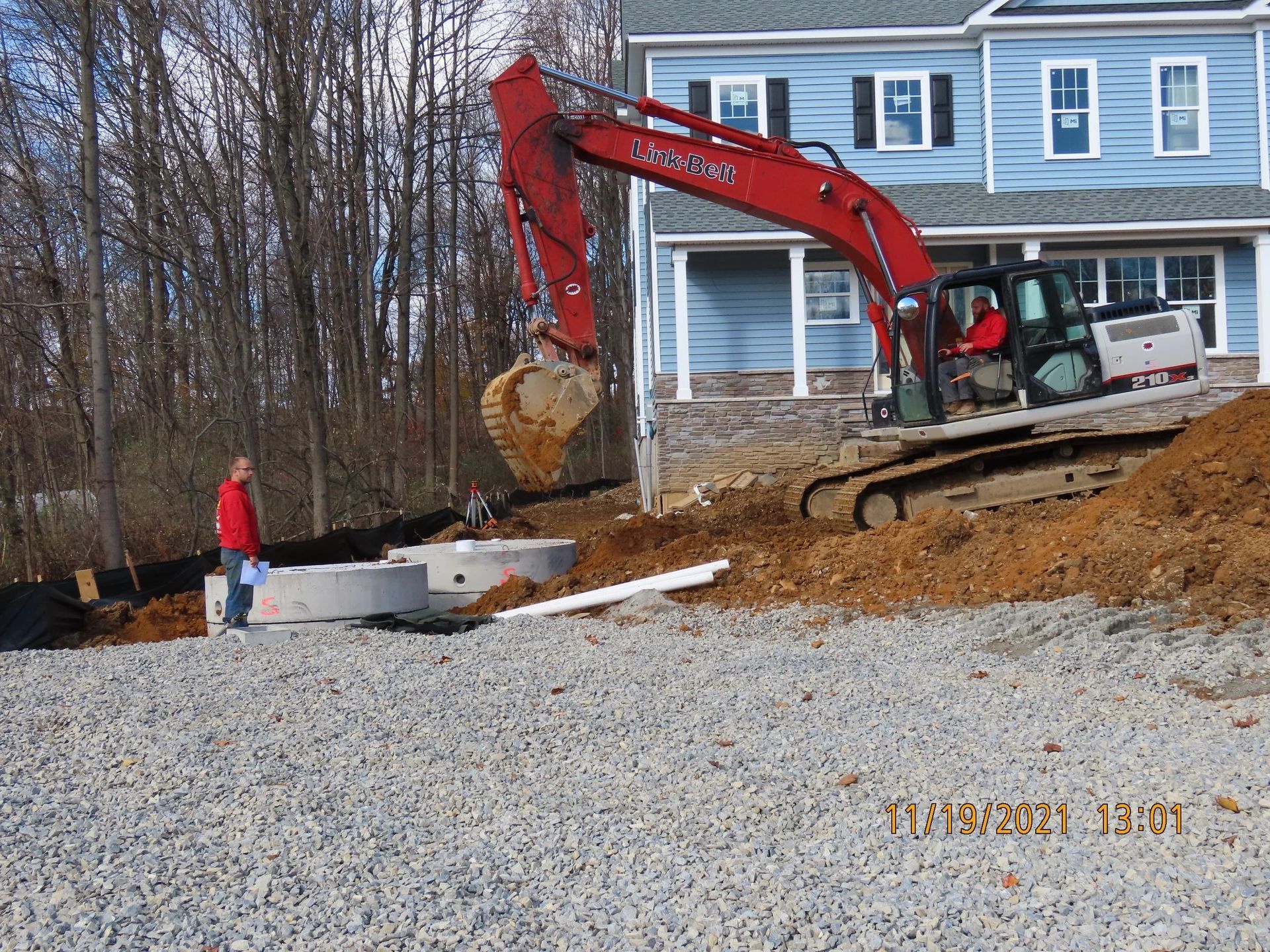 Excavator digging near a light blue house. A person in red watches by concrete structures.