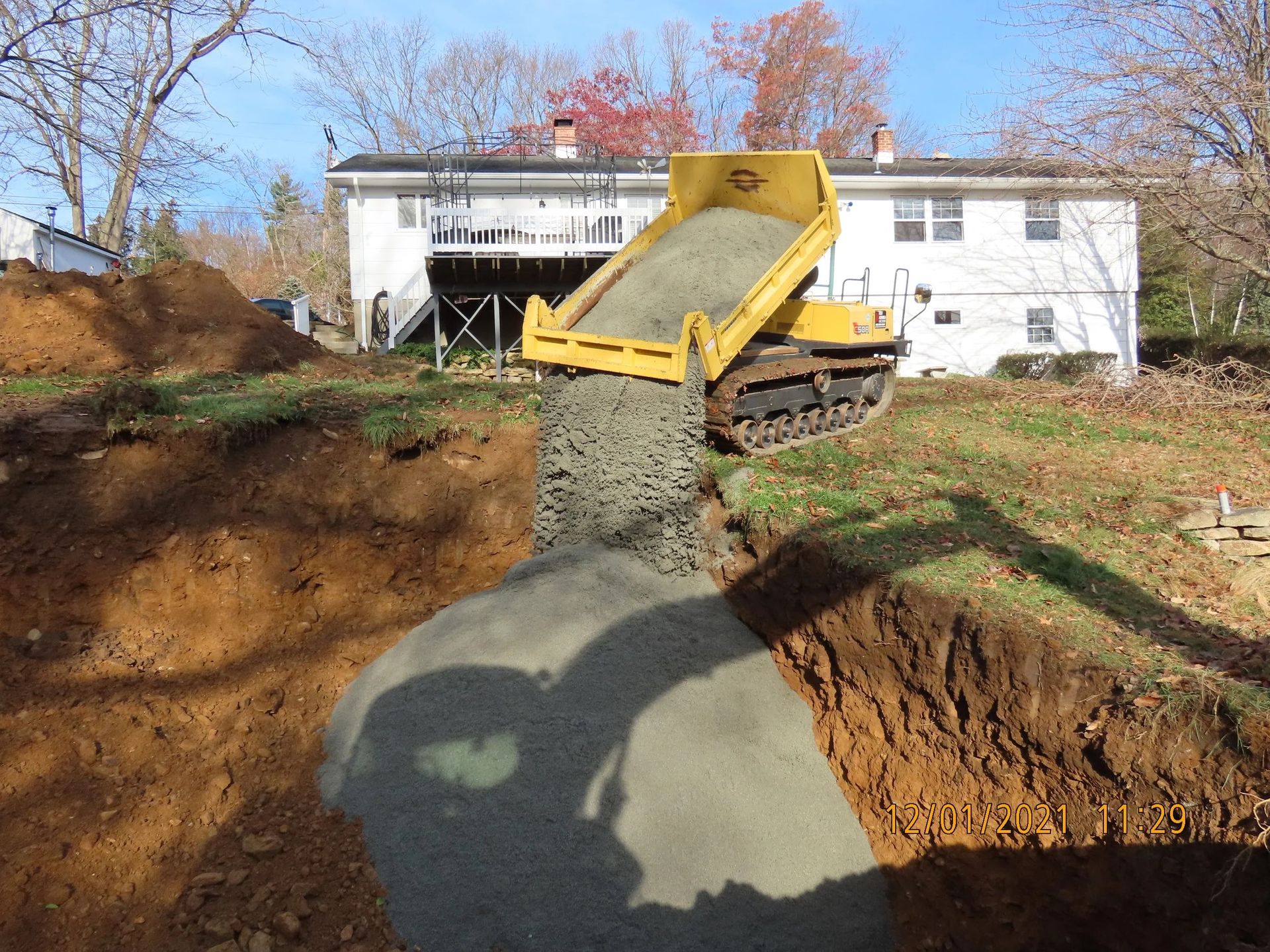 Yellow tracked dump truck dumping gravel into a trench on a residential property.