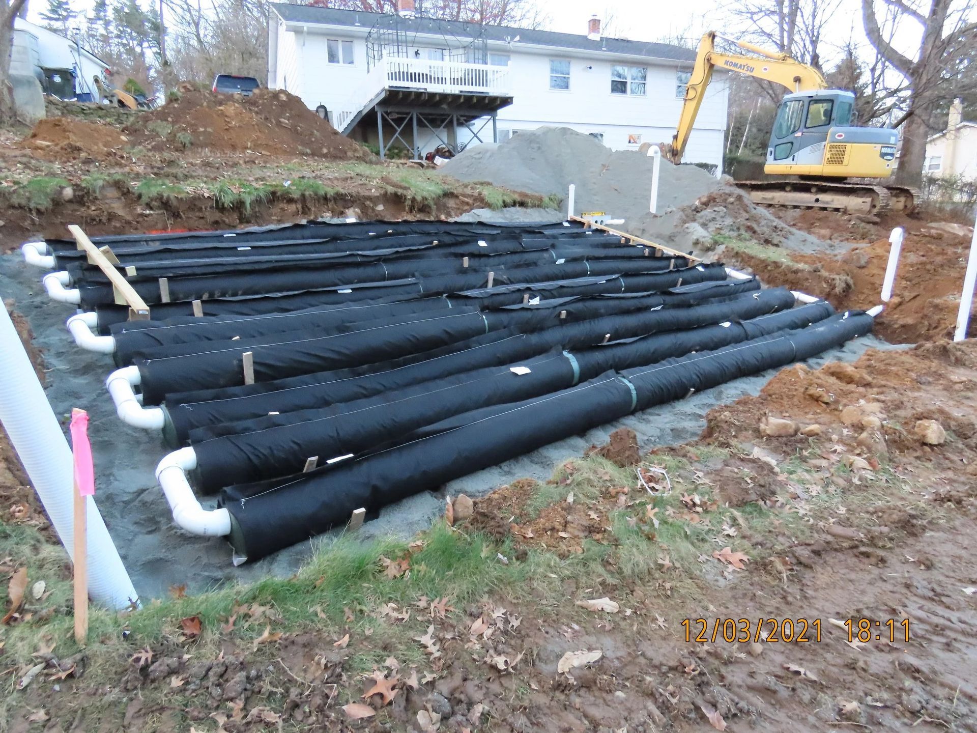 Black drainage tubes installed in a trench, with an excavator in the background.
