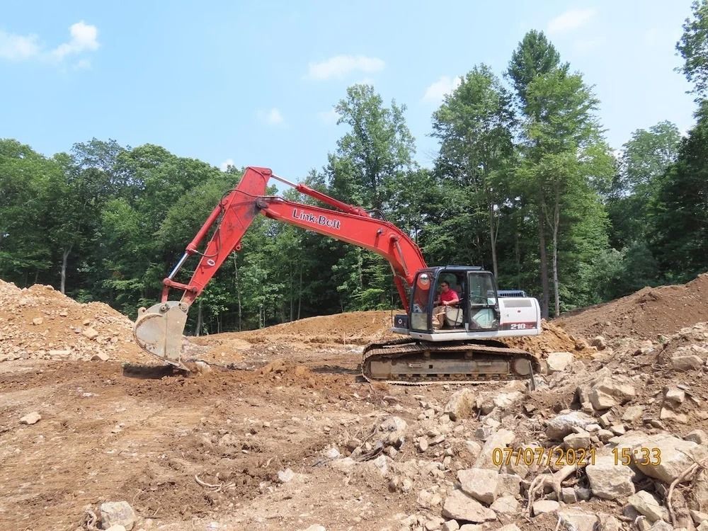 Red excavator working on a dirt lot surrounded by rocks and trees.