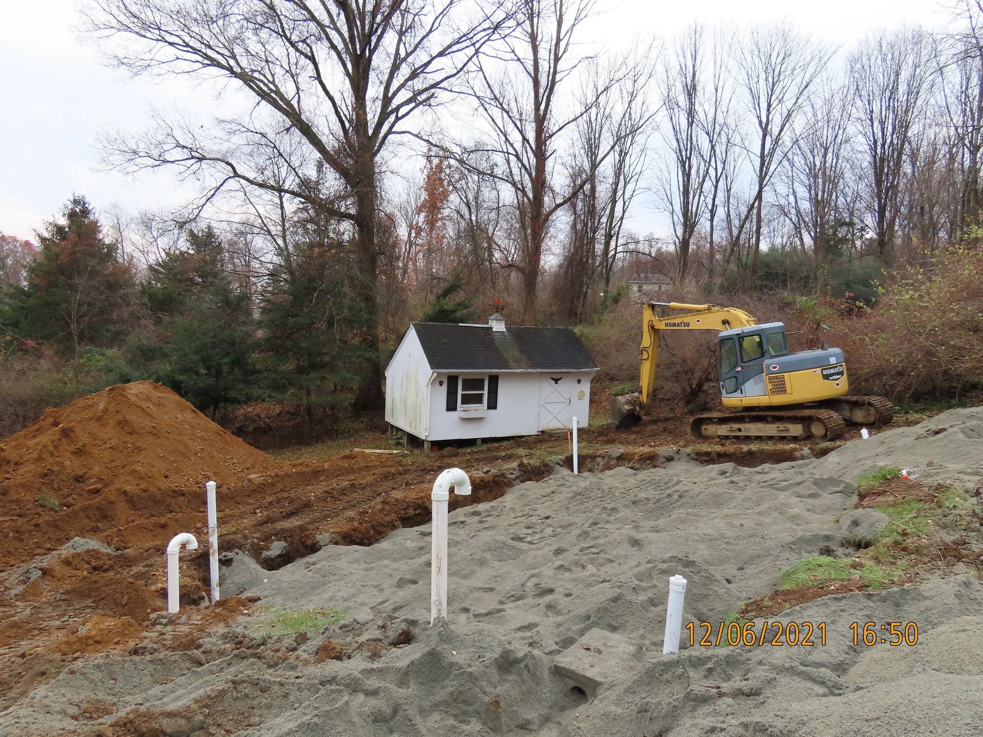 Construction site with a small white building, excavator, and piping in front of a hill with trees.
