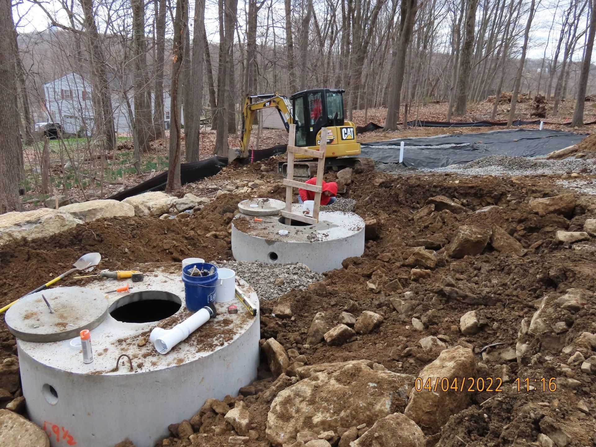 Two concrete septic tanks being installed near a wooded area with a small excavator.