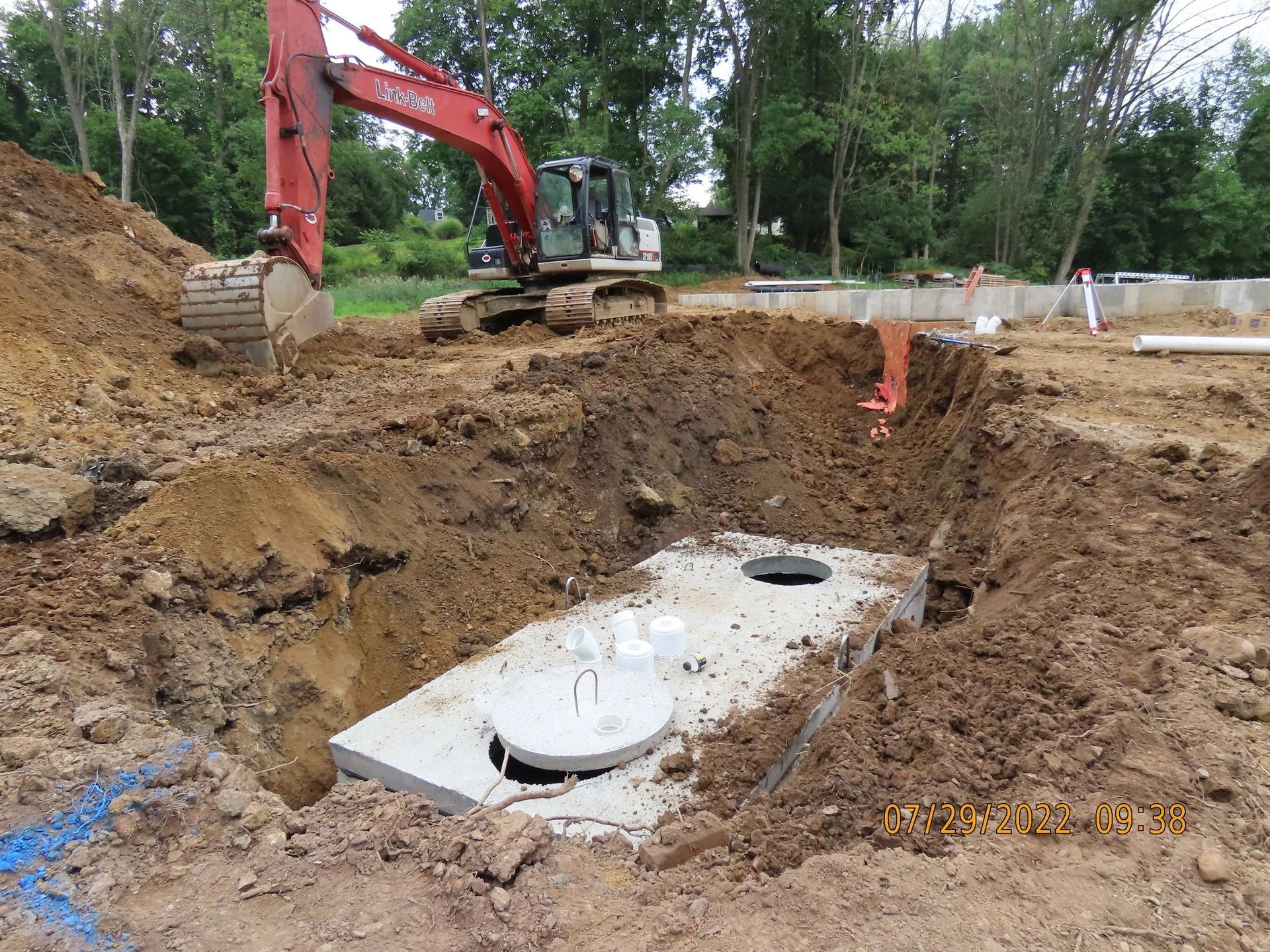 Excavator lowering a concrete septic tank into a dirt hole at a construction site.