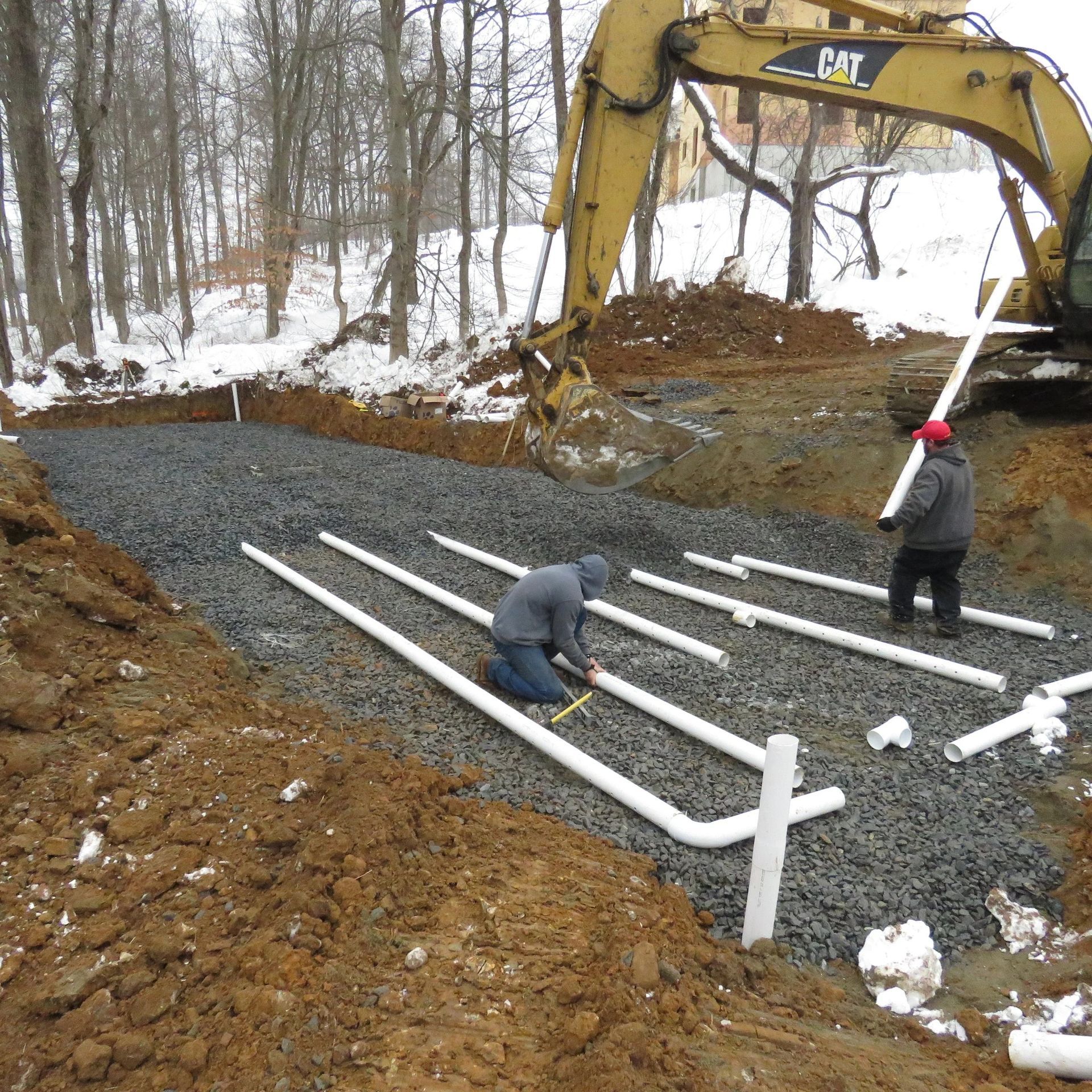 Construction of a septic system; workers install white pipes in a gravel-filled trench with a backhoe in the background.