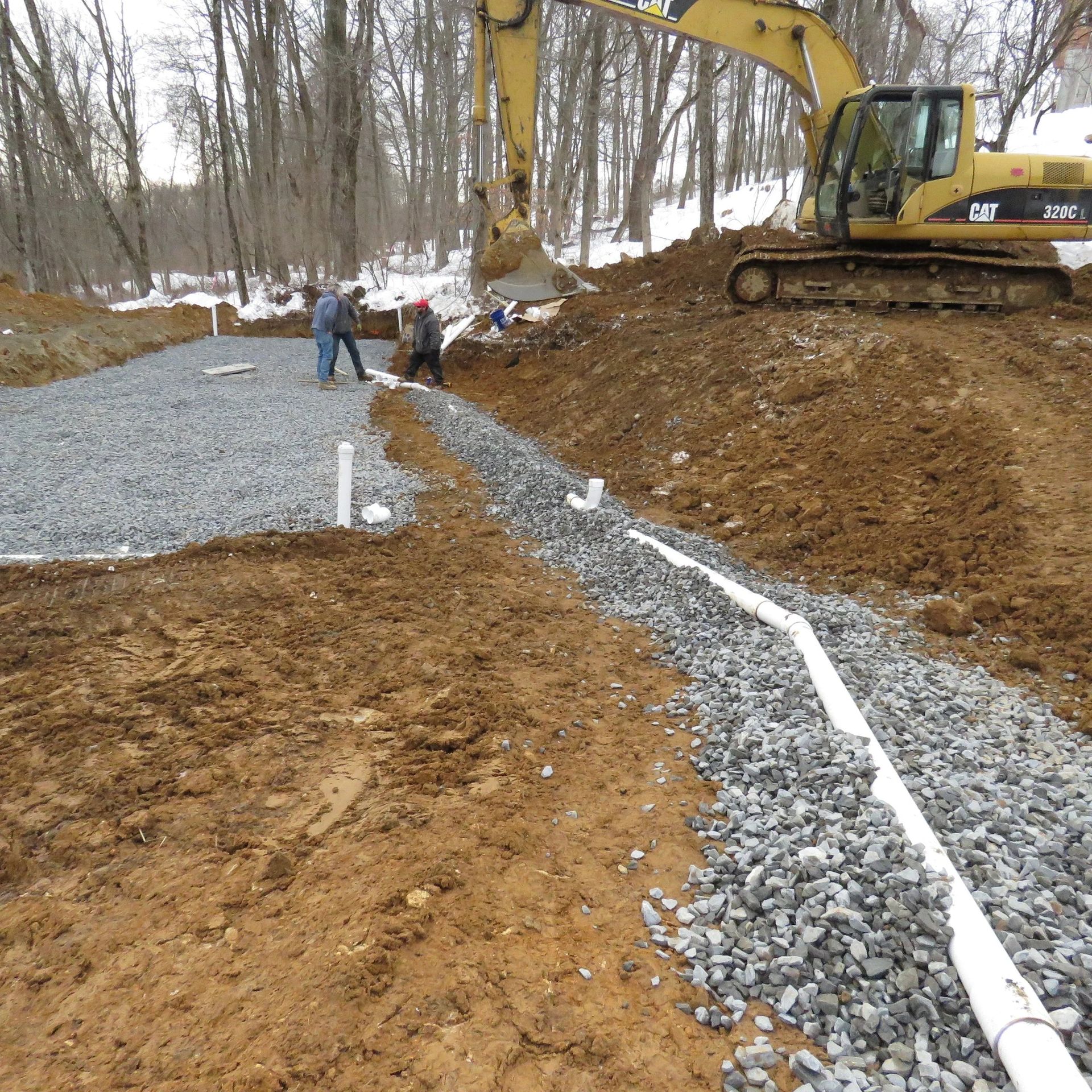 Construction site with an excavator and workers laying pipes and gravel for drainage.